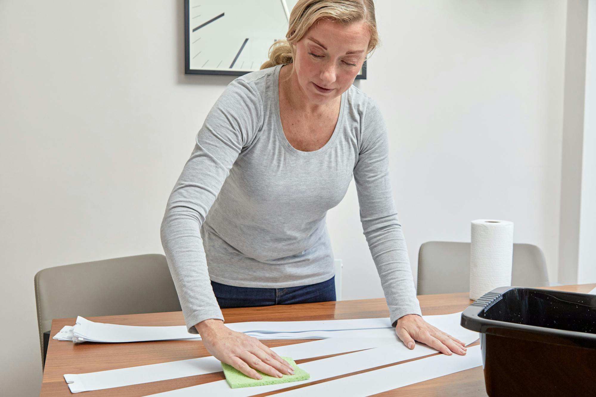 A person wipes a wooden table with a green sponge, surrounded by white paper strips, a roll of paper towels, and a black container in a well-lit room.