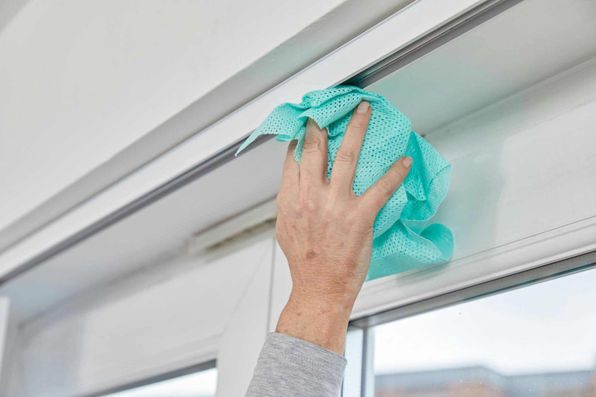 A hand wipes a window frame with a turquoise cloth in a bright room, focusing on cleaning the upper section.