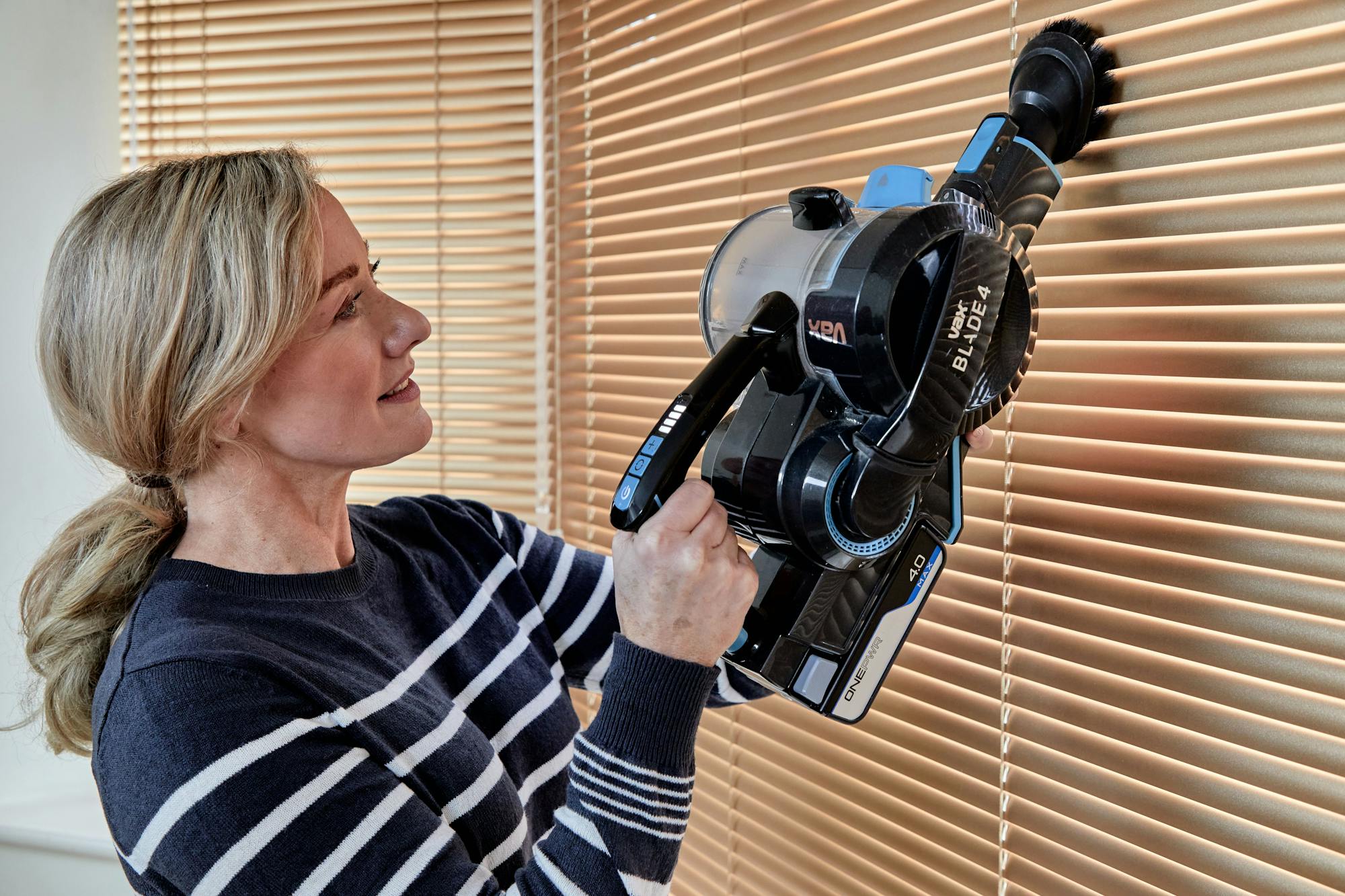 A person uses a "VAX BLADE 4" vacuum cleaner to dust window blinds in a room. The vacuum is handheld, with visible control buttons and a brush attachment.