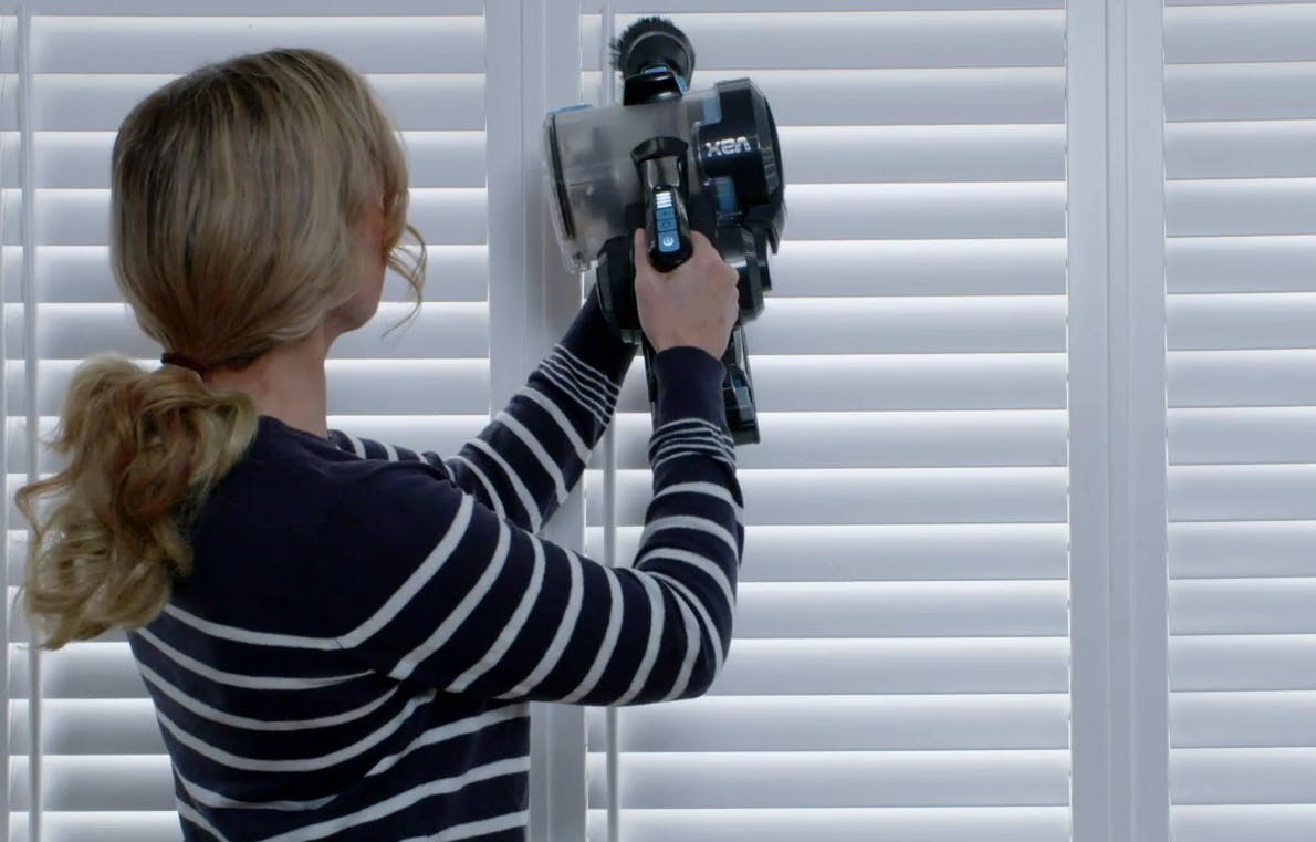 A person is using a VAX cordless vacuum cleaner to clean white window blinds. They're wearing a black and white striped shirt in a bright indoor setting.