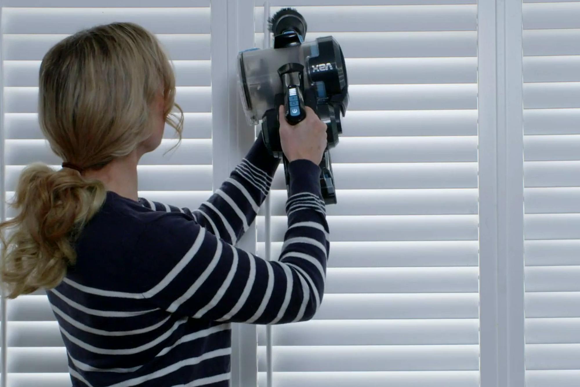 A person is using a VAX cordless vacuum cleaner to clean white window blinds. They're wearing a black and white striped shirt in a bright indoor setting.