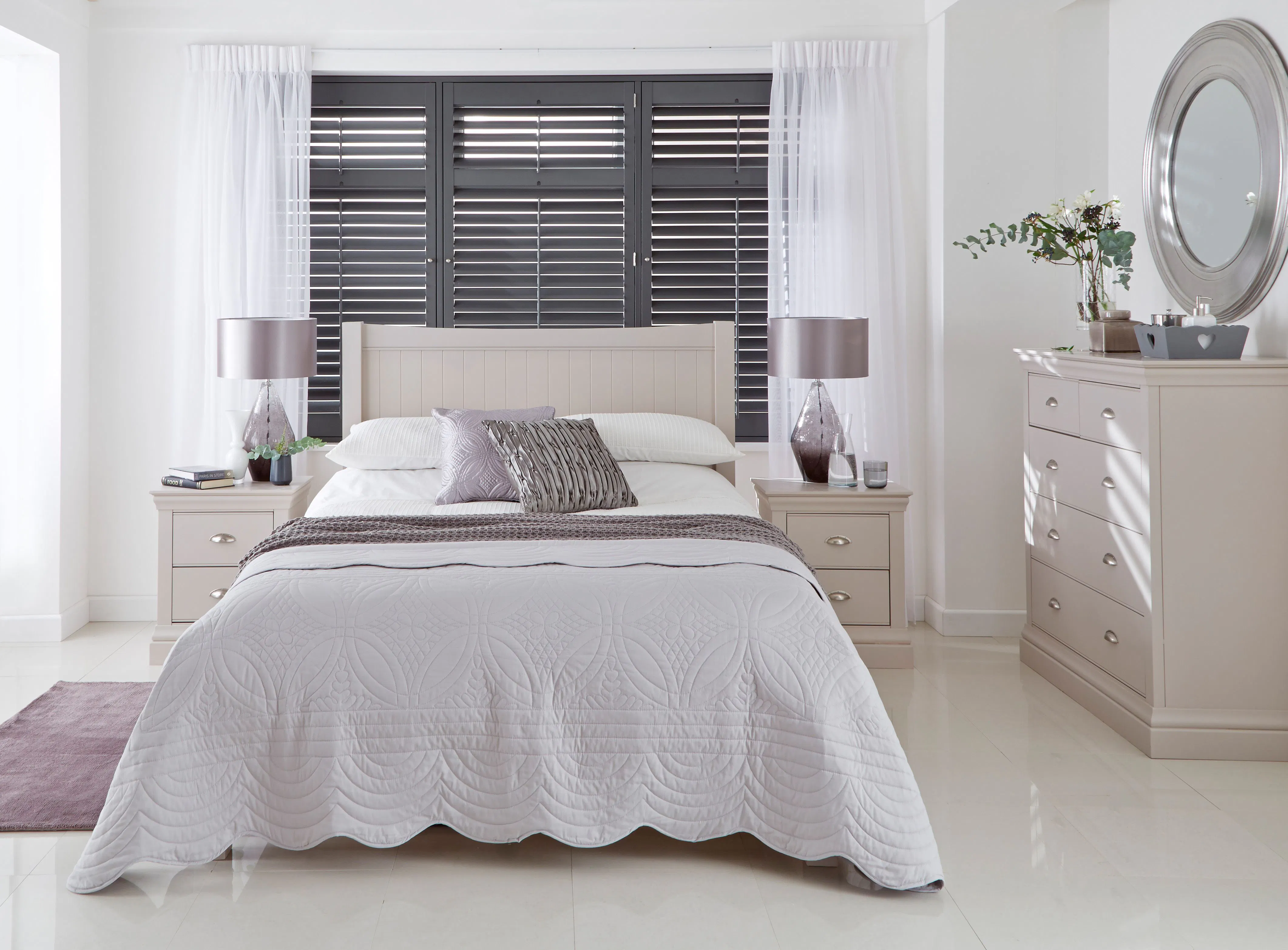 A neatly made bed with decorative pillows sits against a wall with dark shutters. Flanked by matching nightstands with lamps, it faces a dresser with a round mirror.