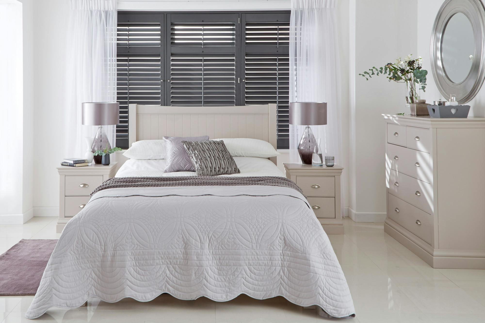 A neatly made bed with decorative pillows sits against a wall with dark shutters. Flanked by matching nightstands with lamps, it faces a dresser with a round mirror.