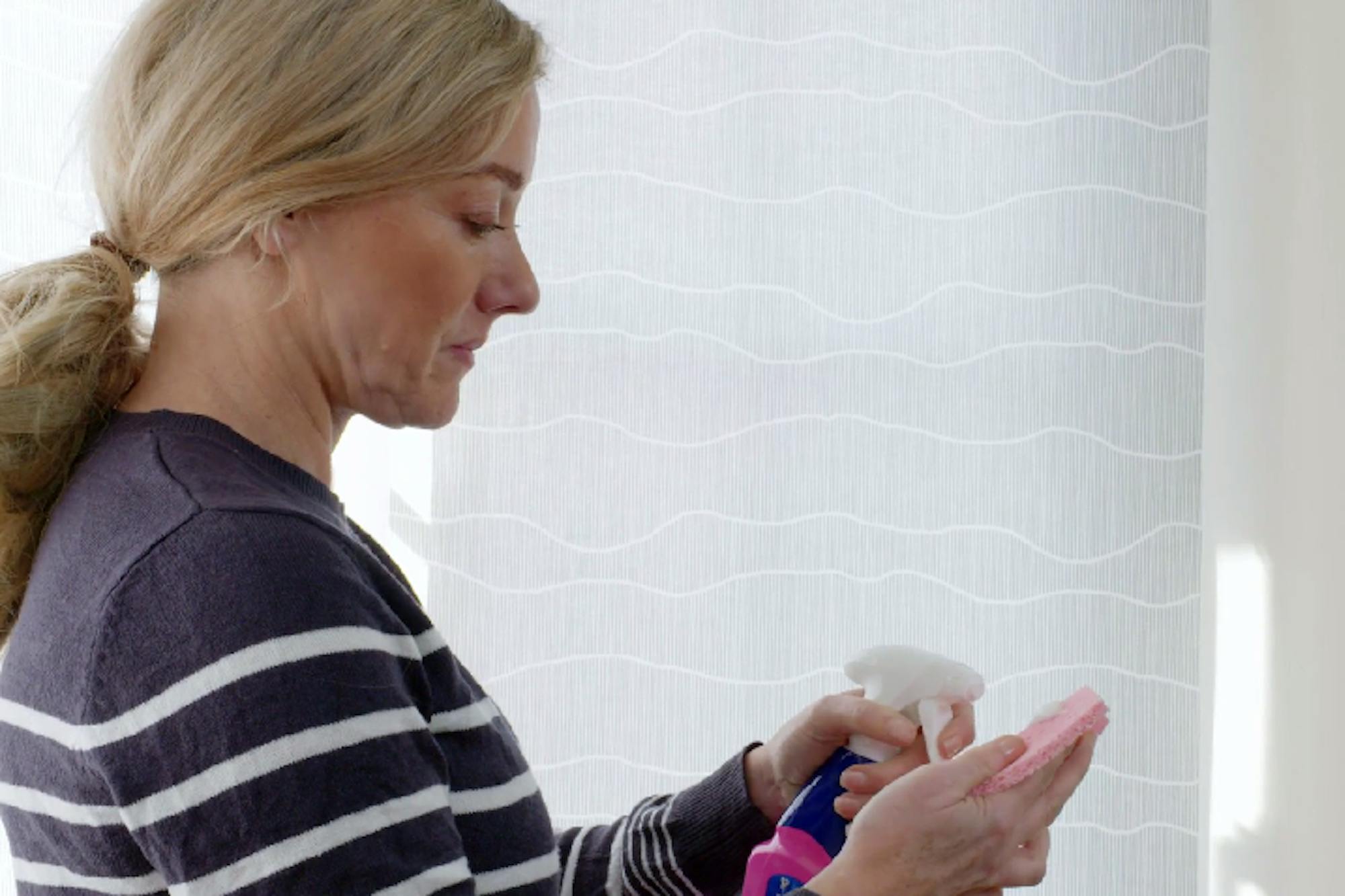 A person with blonde hair, wearing a striped sweater, sprays cleaner onto a cloth. They are indoors, standing in front of a window with a wavy patterned curtain.