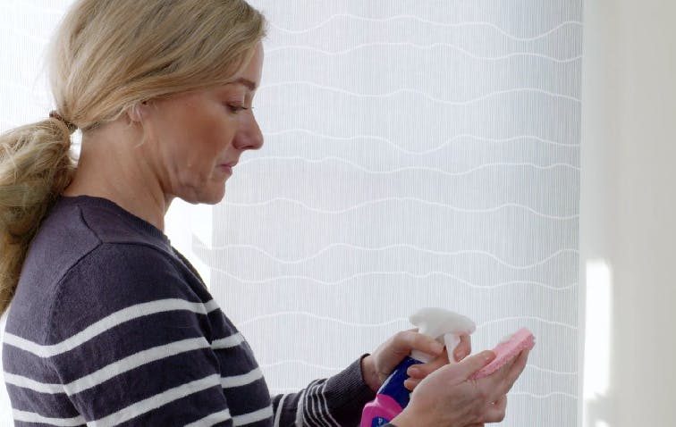 A person with blonde hair, wearing a striped sweater, sprays cleaner onto a cloth. They are indoors, standing in front of a window with a wavy patterned curtain.