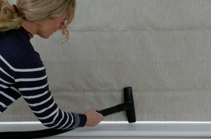 A person in a striped sweater uses a vacuum cleaner attachment to clean a fabric-covered wall. The setting appears to be indoors, with minimal visible furnishings.