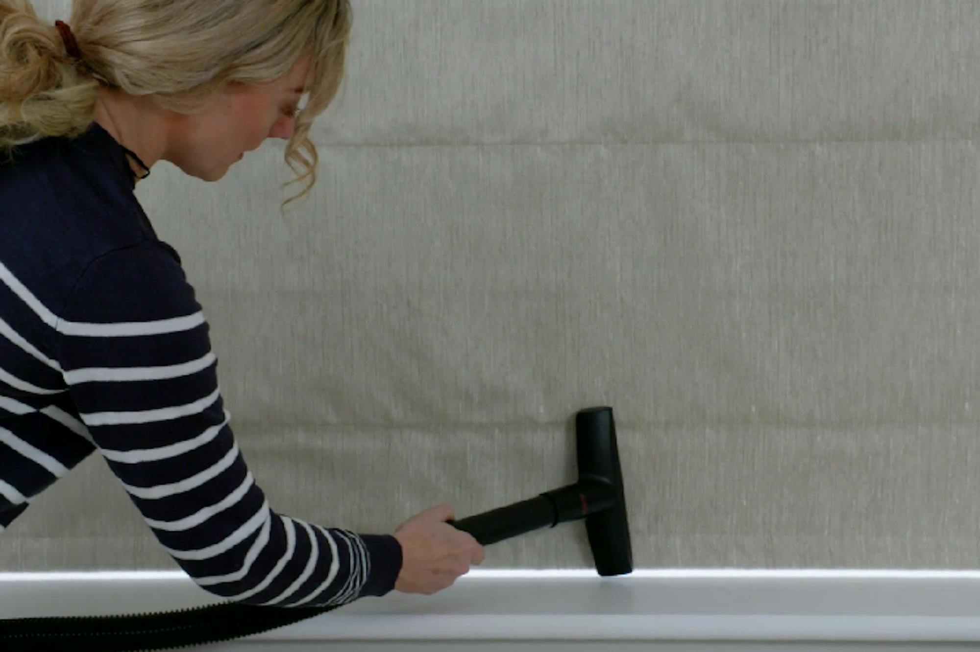 A person in a striped sweater uses a vacuum cleaner attachment to clean a fabric-covered wall. The setting appears to be indoors, with minimal visible furnishings.