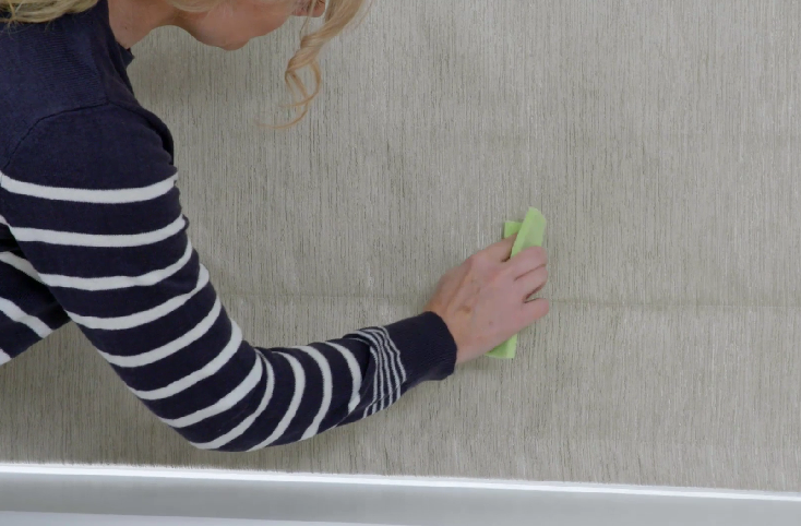 A person in a striped sweater uses a green cloth to clean or smooth a textured, beige wall. The upper body is partially visible, and the setting appears domestic.