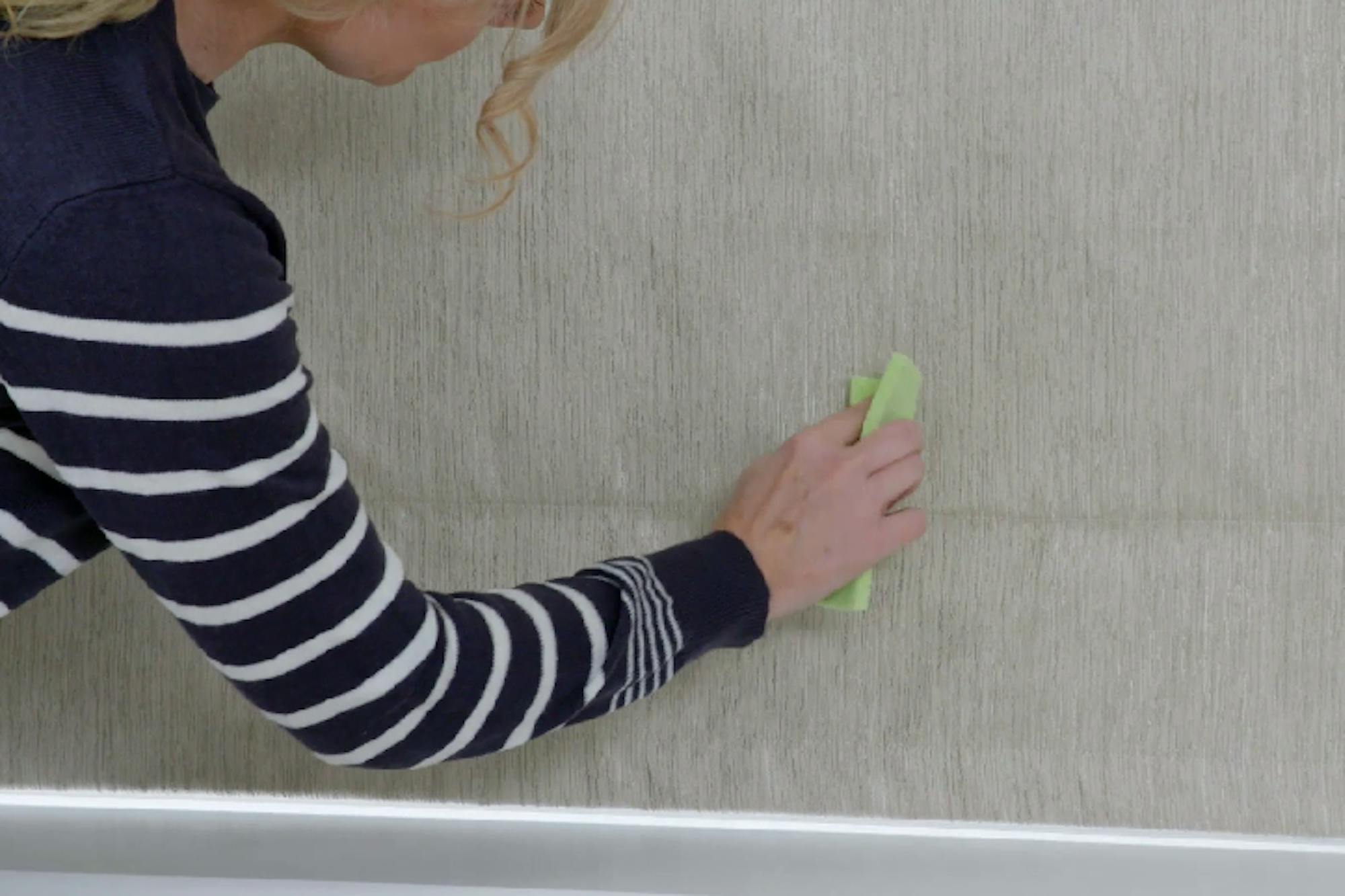 A person in a striped sweater uses a green cloth to clean or smooth a textured, beige wall. The upper body is partially visible, and the setting appears domestic.