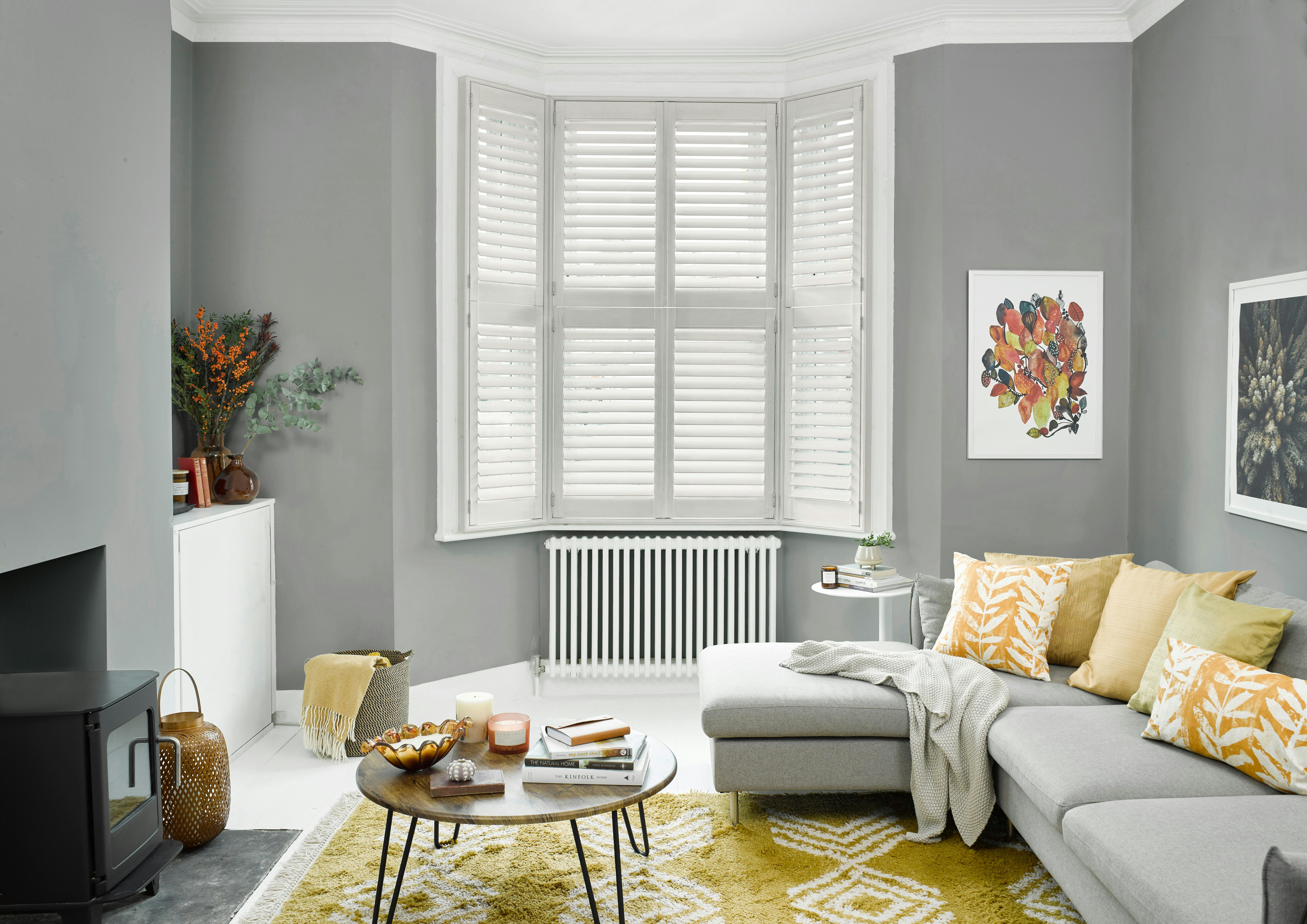 A cozy living room features a gray sectional sofa with patterned cushions. A wooden coffee table with books and candles sits on a yellow rug. White shutters cover bay windows.