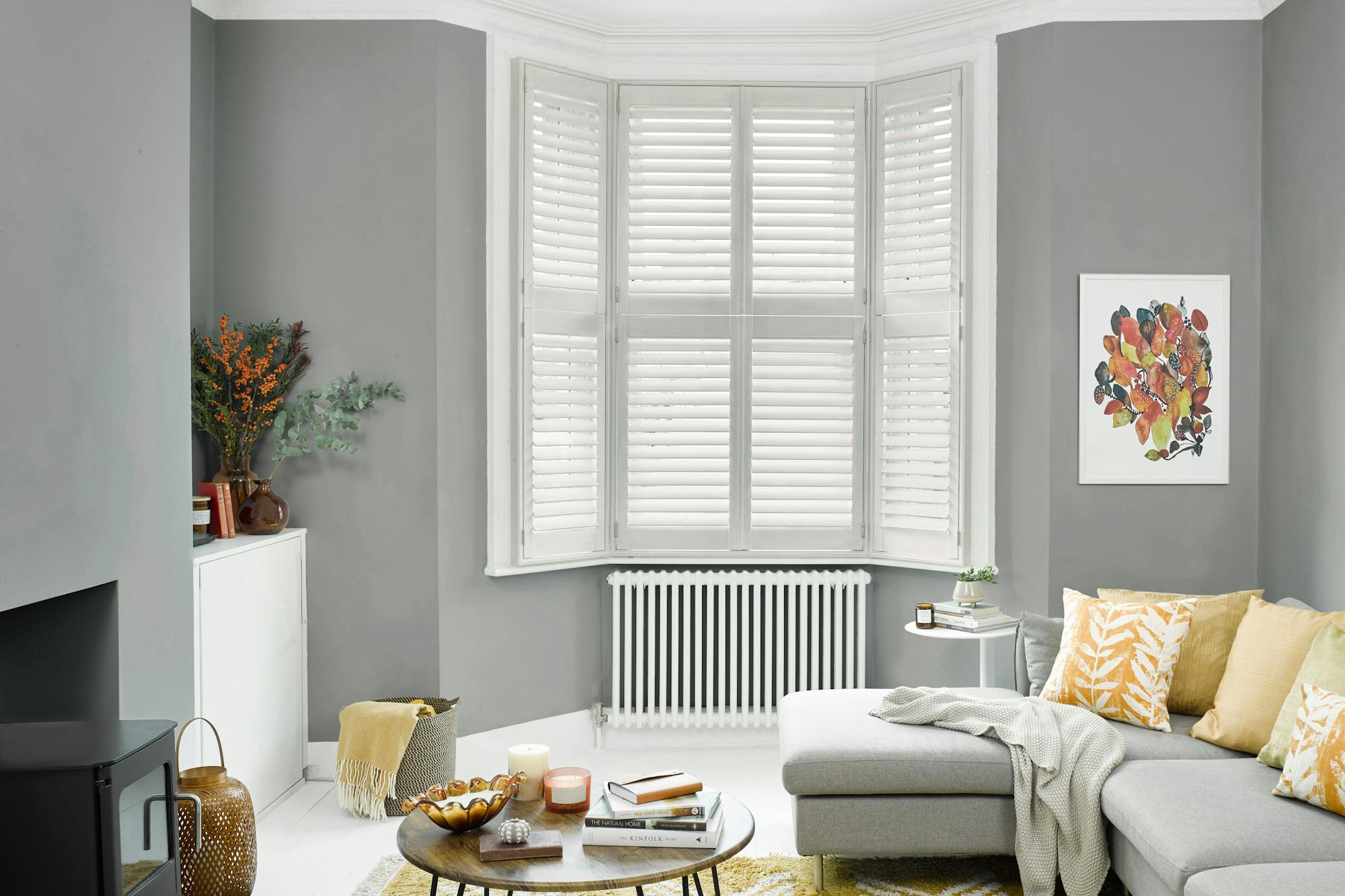 A cozy living room features a gray sectional sofa with patterned cushions. A wooden coffee table with books and candles sits on a yellow rug. White shutters cover bay windows.