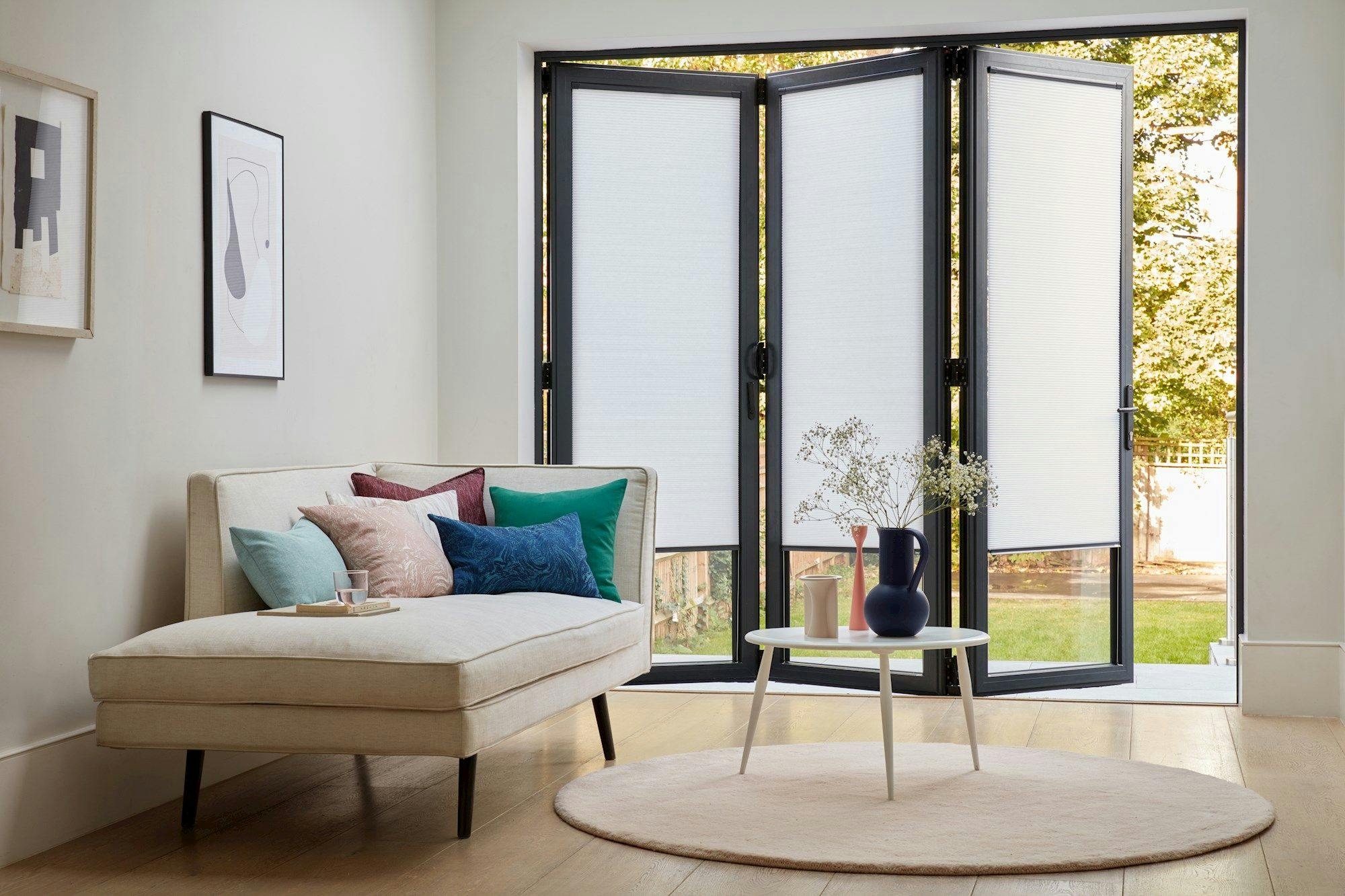 A beige chaise lounge with colorful pillows rests in a bright room. Nearby, a round table holds a vase with branches, set against large folding patio doors opening to a garden.