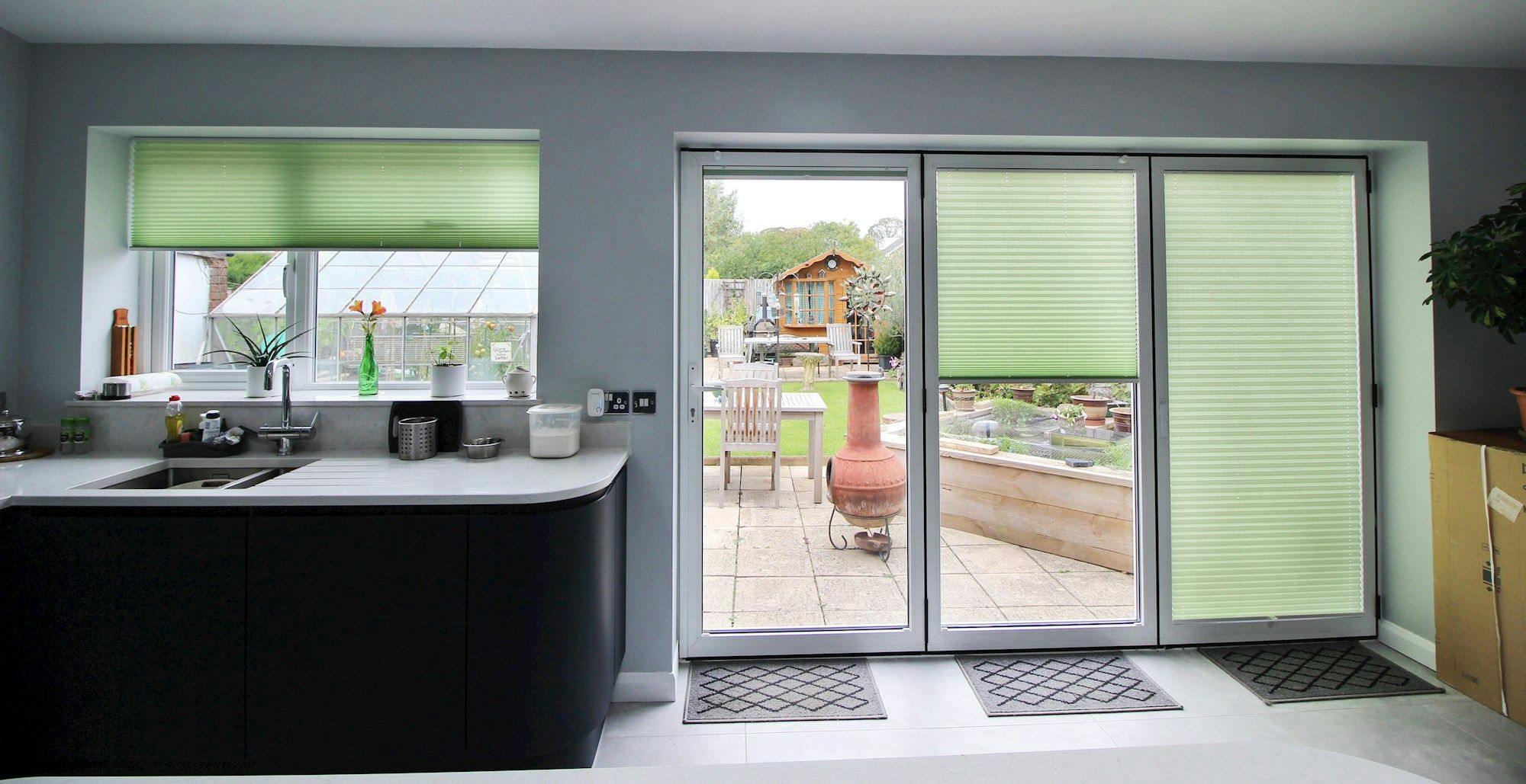 Kitchen counter with a sink and plants sits under a window with green blinds. Sliding glass doors open to a backyard with patio furniture and a wooden shed.