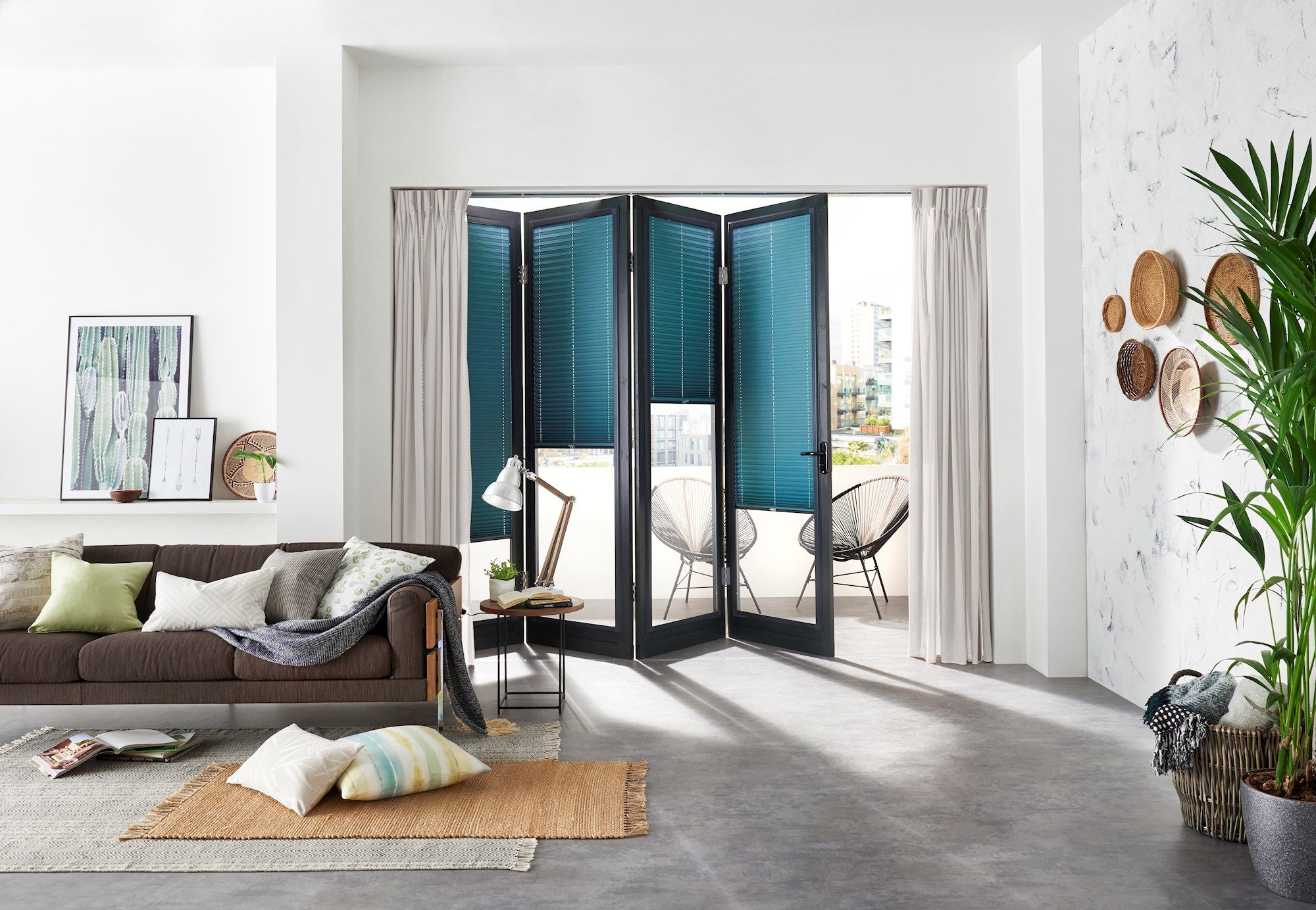 Sofa adorned with pillows and a blanket, sitting next to a side table with a lamp in a bright, modern living room. Large bifold doors open to a balcony with chairs.