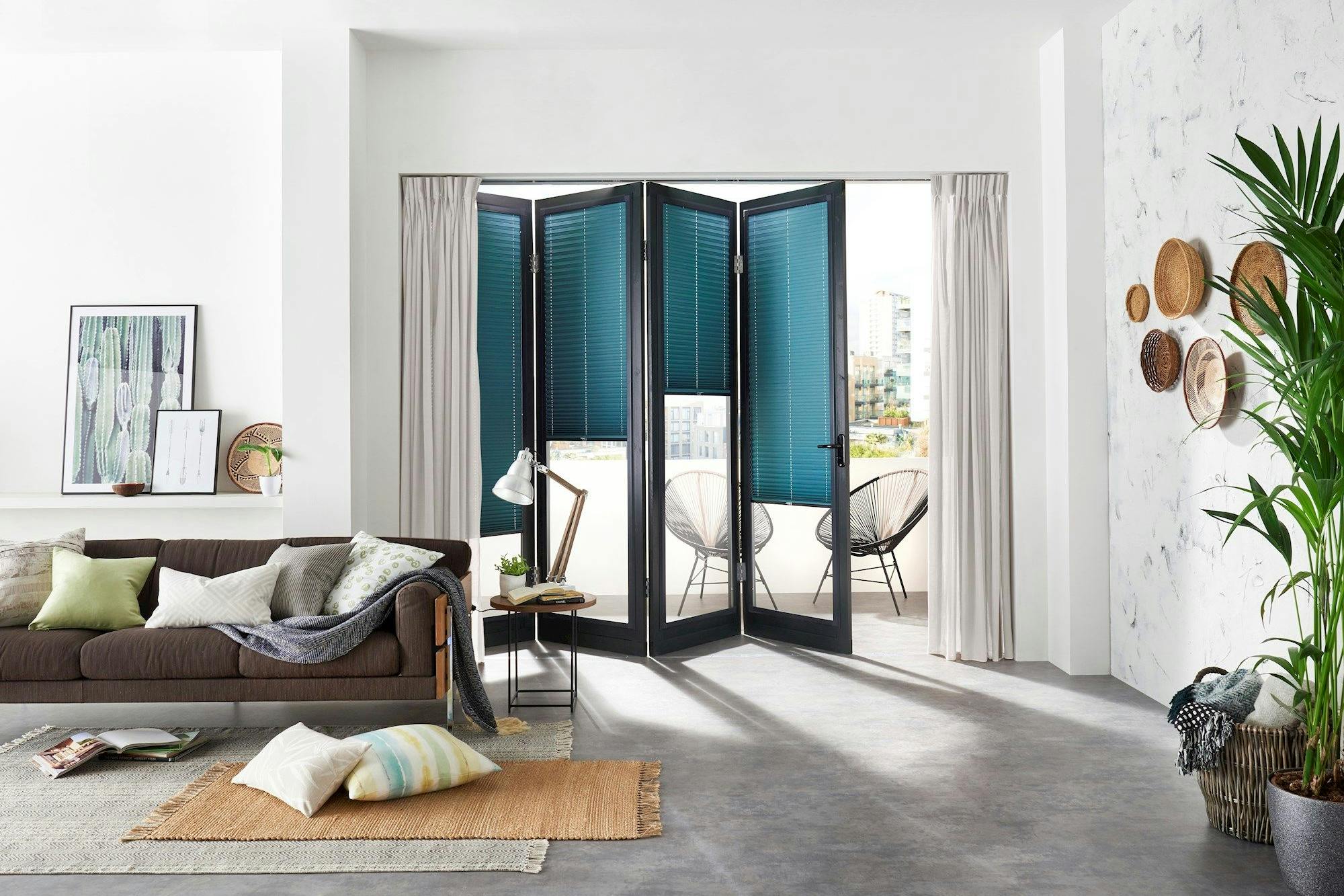 Sofa adorned with pillows and a blanket, sitting next to a side table with a lamp in a bright, modern living room. Large bifold doors open to a balcony with chairs.