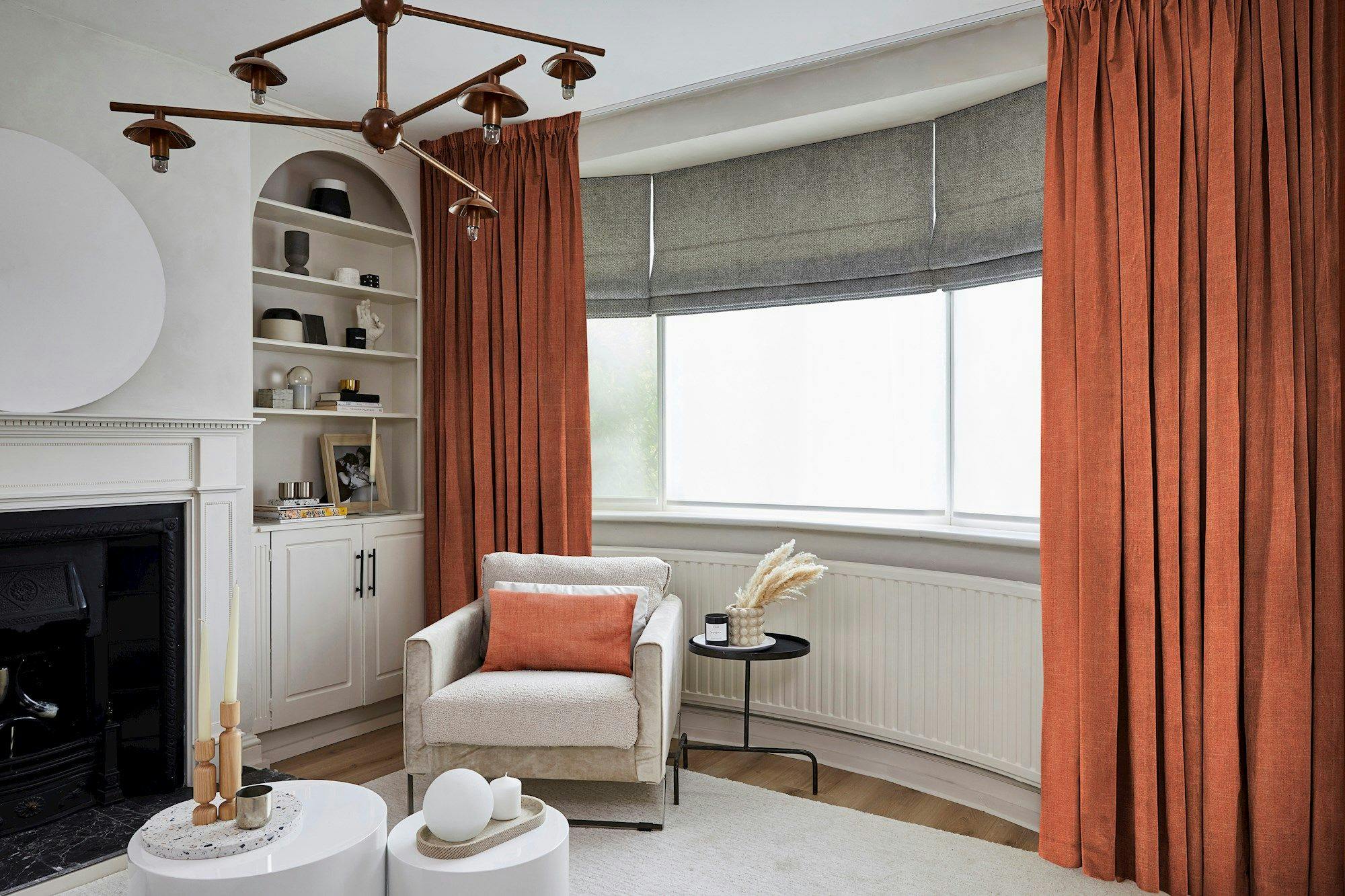 Armchair placed near a window with orange curtains in a cozy living room; features a modern chandelier, built-in shelves with decor, round tables, and a black fireplace.
