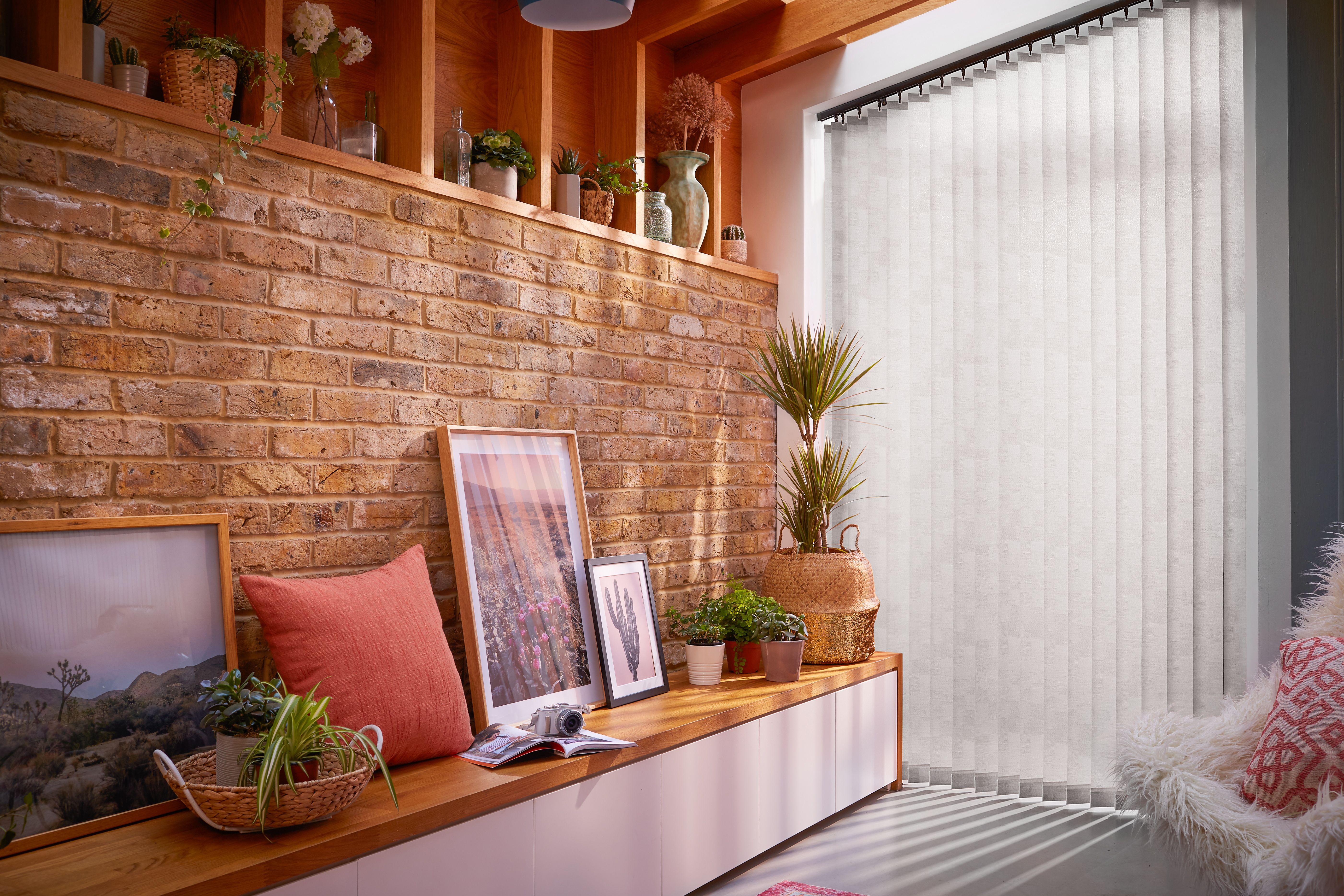 Framed photographs and a red pillow rest on a wooden bench against a brick wall. Various plants decorate the space. Vertical blinds cover a window, softly filtering daylight.