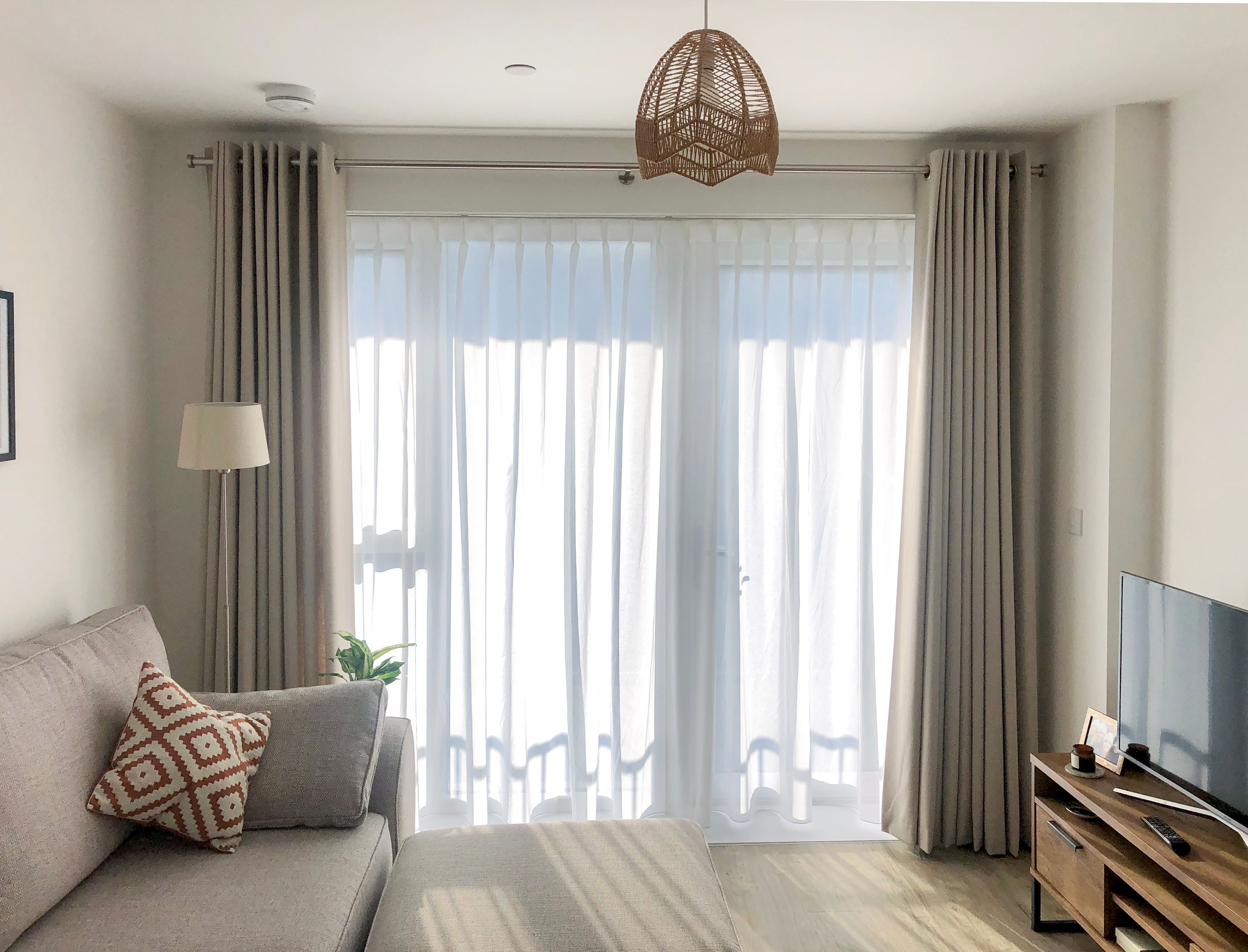 A cozy living room features a gray sofa with patterned cushions, placed next to a lamp and plant. Sunlight filters through sheer curtains, illuminating the TV and wooden stand.