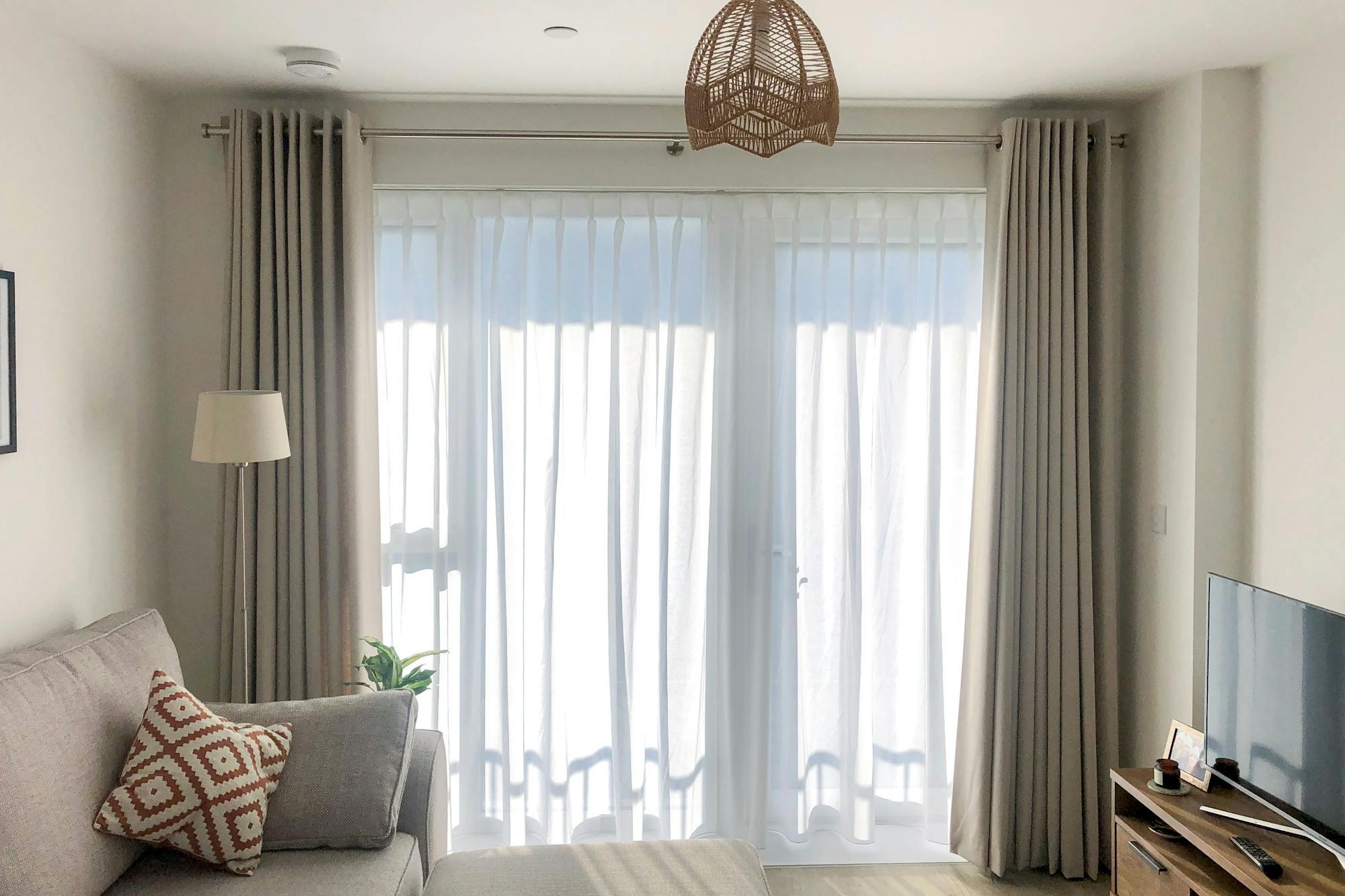 A cozy living room features a gray sofa with patterned cushions, placed next to a lamp and plant. Sunlight filters through sheer curtains, illuminating the TV and wooden stand.