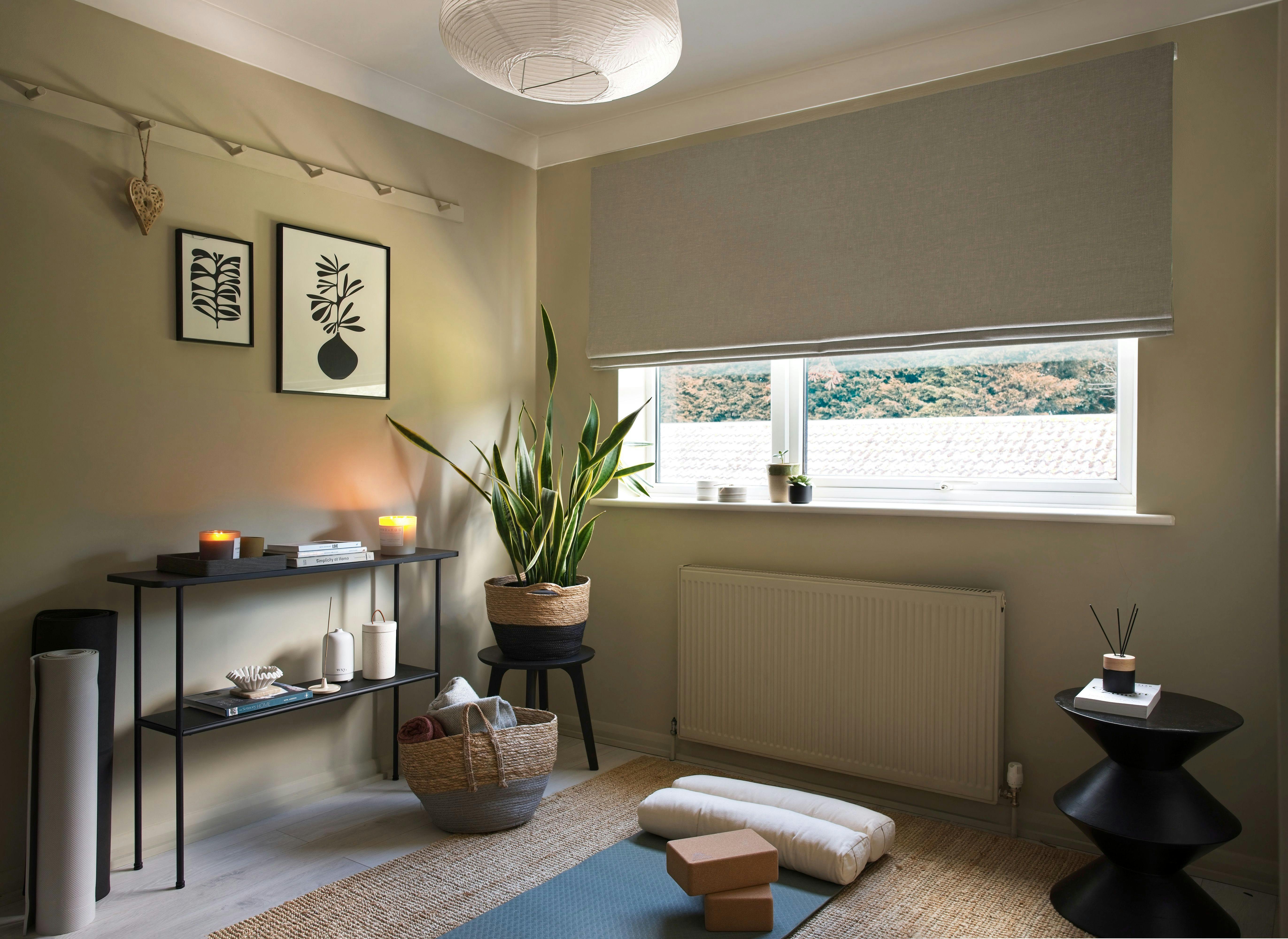 Yoga mat lies in a serene room with rolled towels and cork blocks, surrounded by plants, candles, artwork, and a window with natural light illuminating the space.