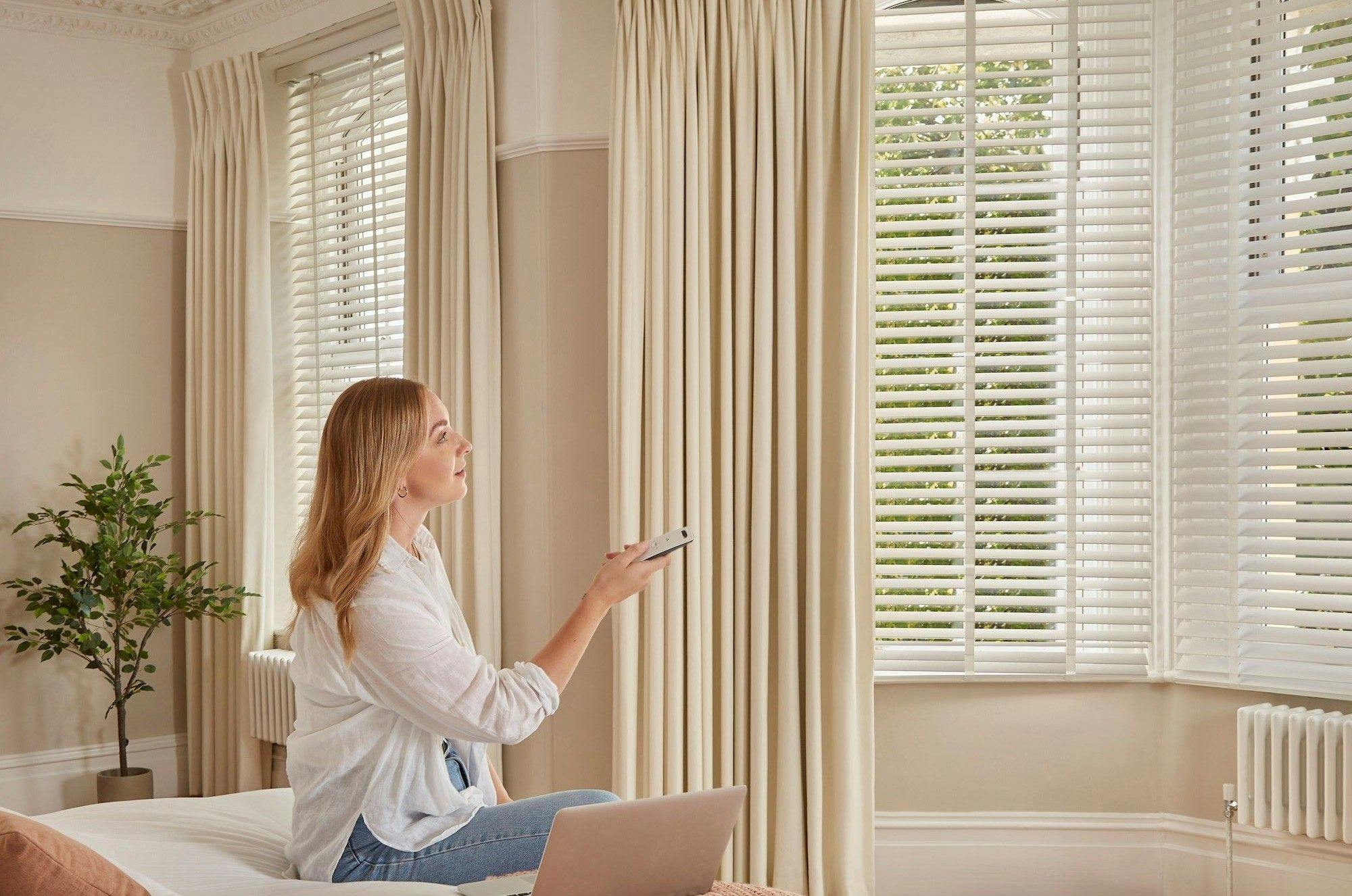 A woman holds a remote control, pointing it towards automated blinds. She sits on a bed in a bright, elegantly furnished room with cream curtains and a potted plant.