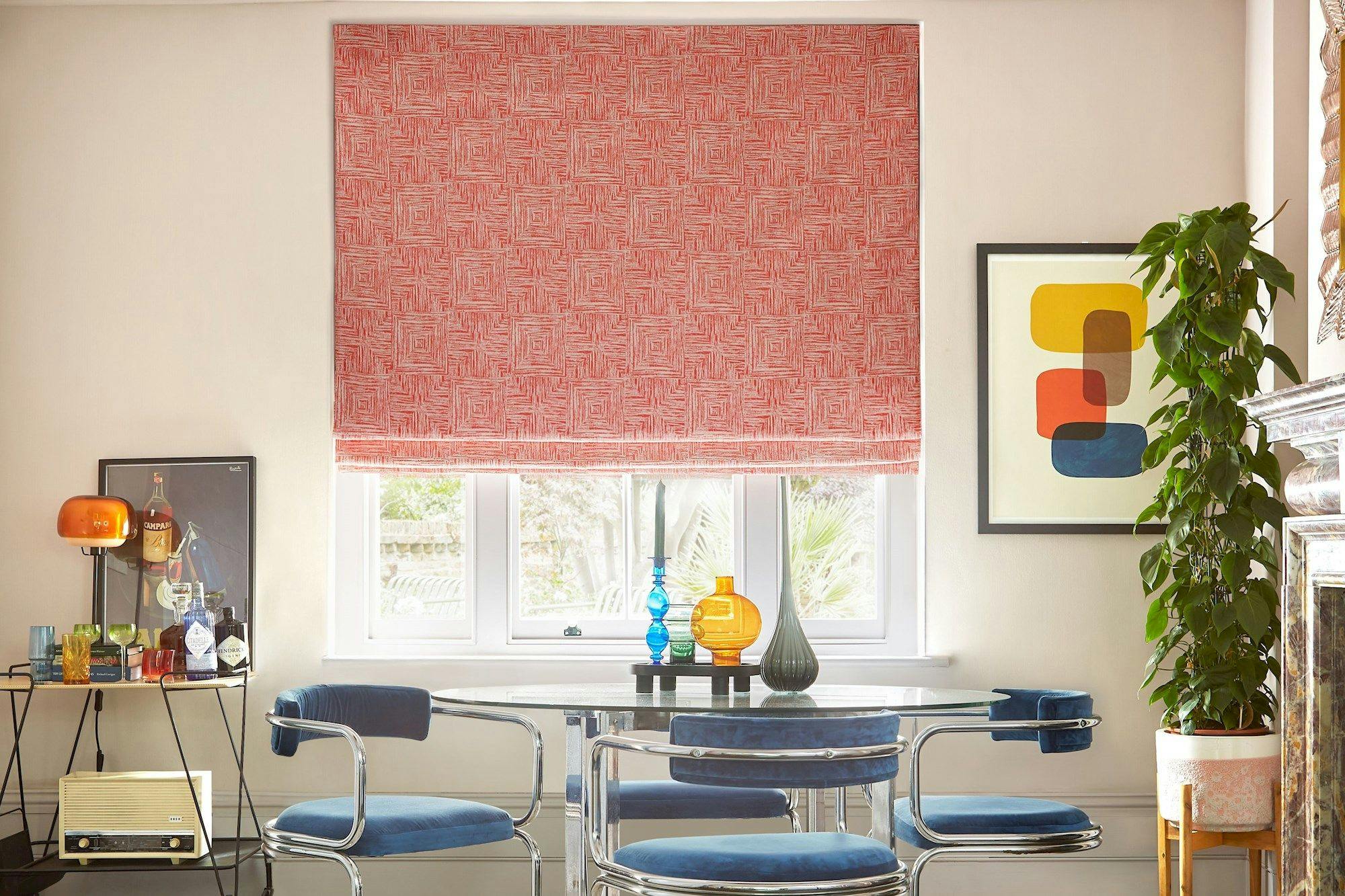 A patterned red blind covers a window behind a modern dining table with blue chairs. A side table holds glassware and lamps. Colorful abstract art and a plant decorate the room.