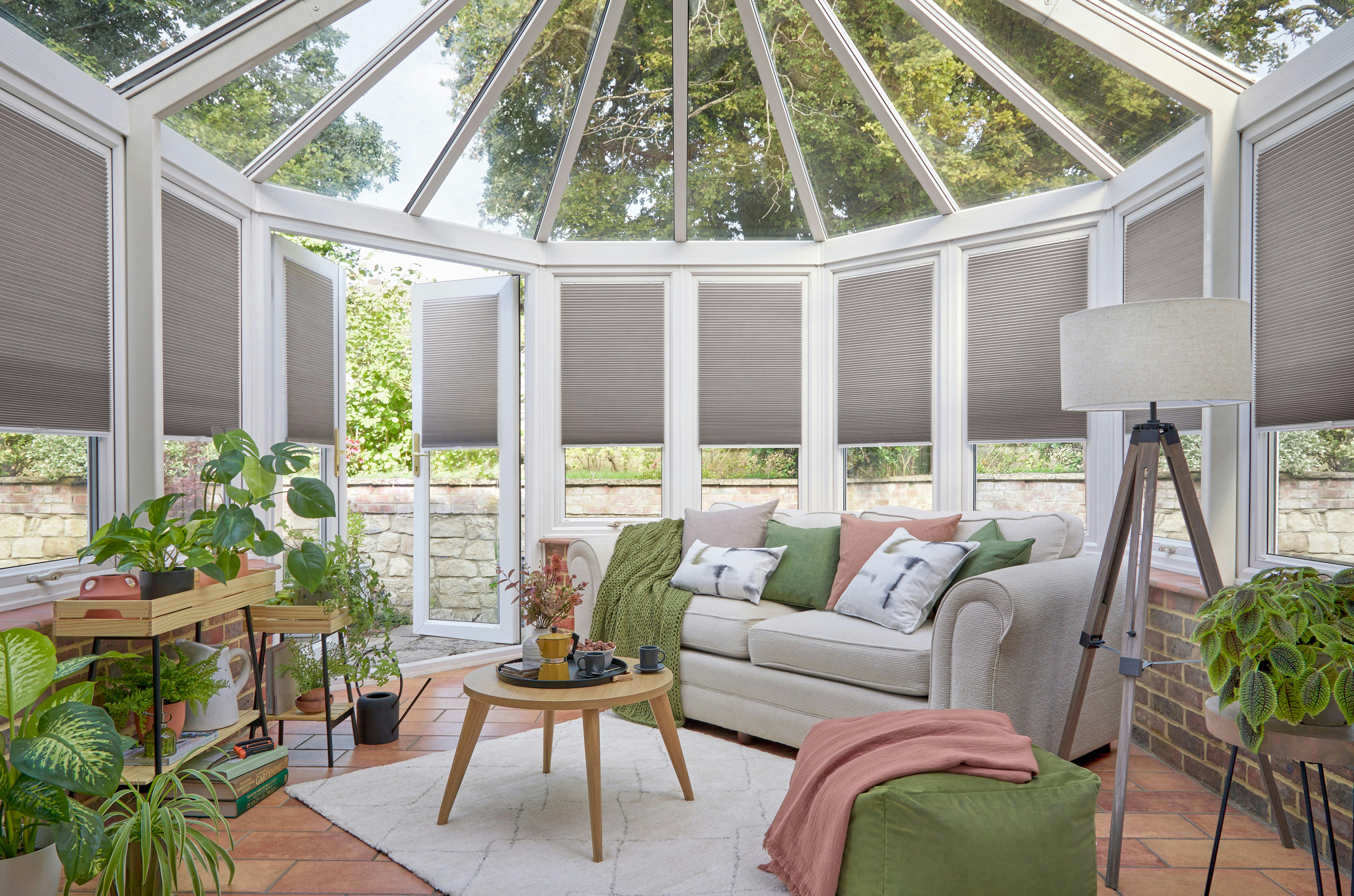 A cozy conservatory features a beige sofa adorned with colorful pillows, surrounded by lush green plants. A wooden coffee table sits on a rug, complemented by an elegant floor lamp.