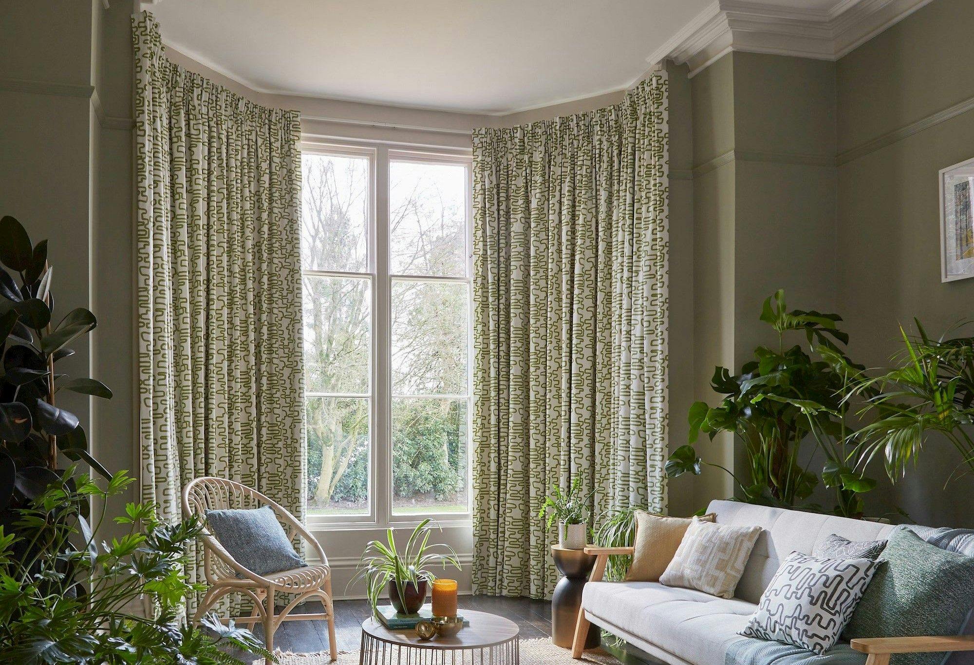 Curtains hang beside a large window in a living room, surrounding a wicker chair and a table with plants. A sofa with cushions is nearby, and plants decorate the space.