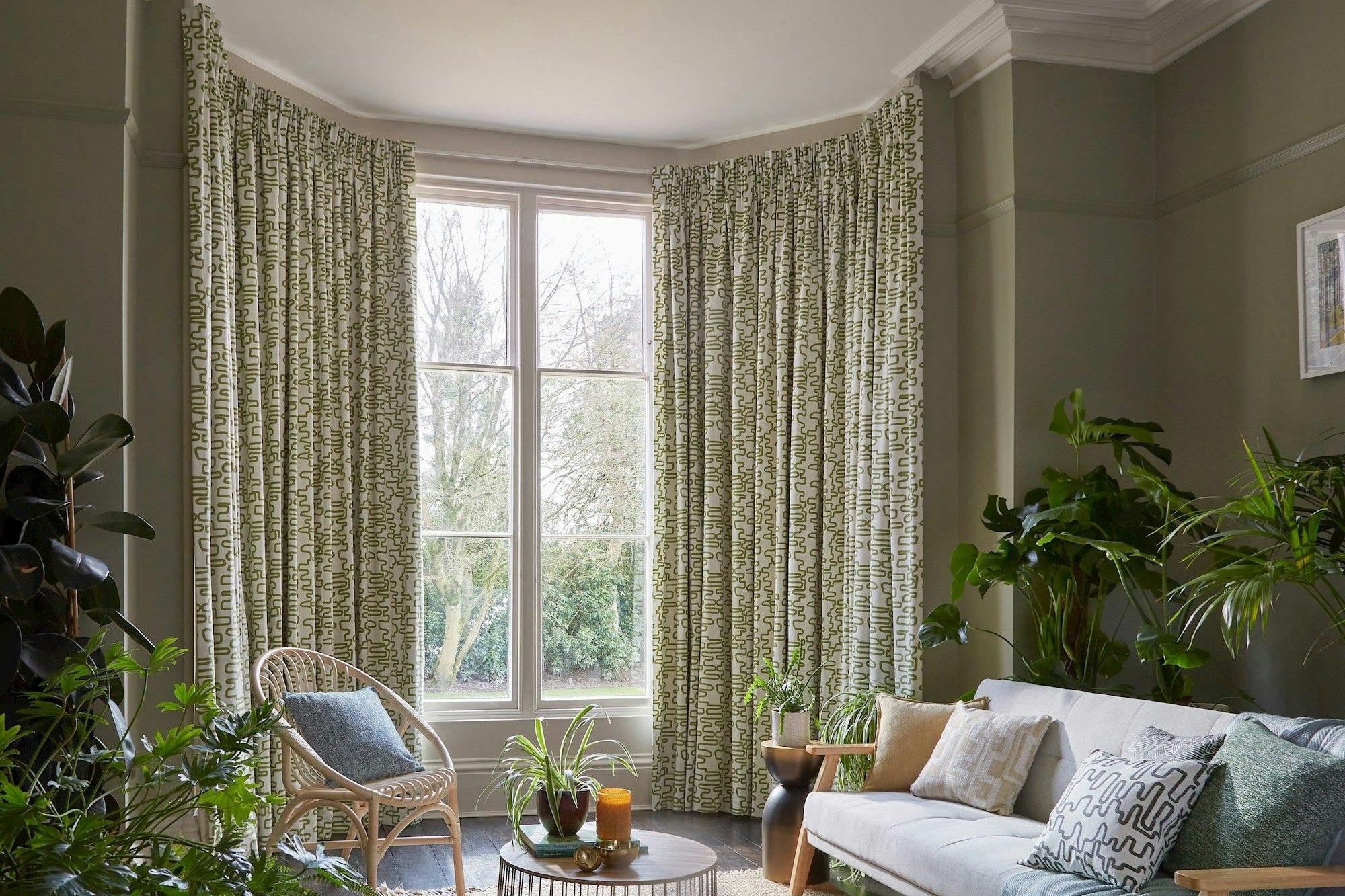Curtains hang beside a large window in a living room, surrounding a wicker chair and a table with plants. A sofa with cushions is nearby, and plants decorate the space.