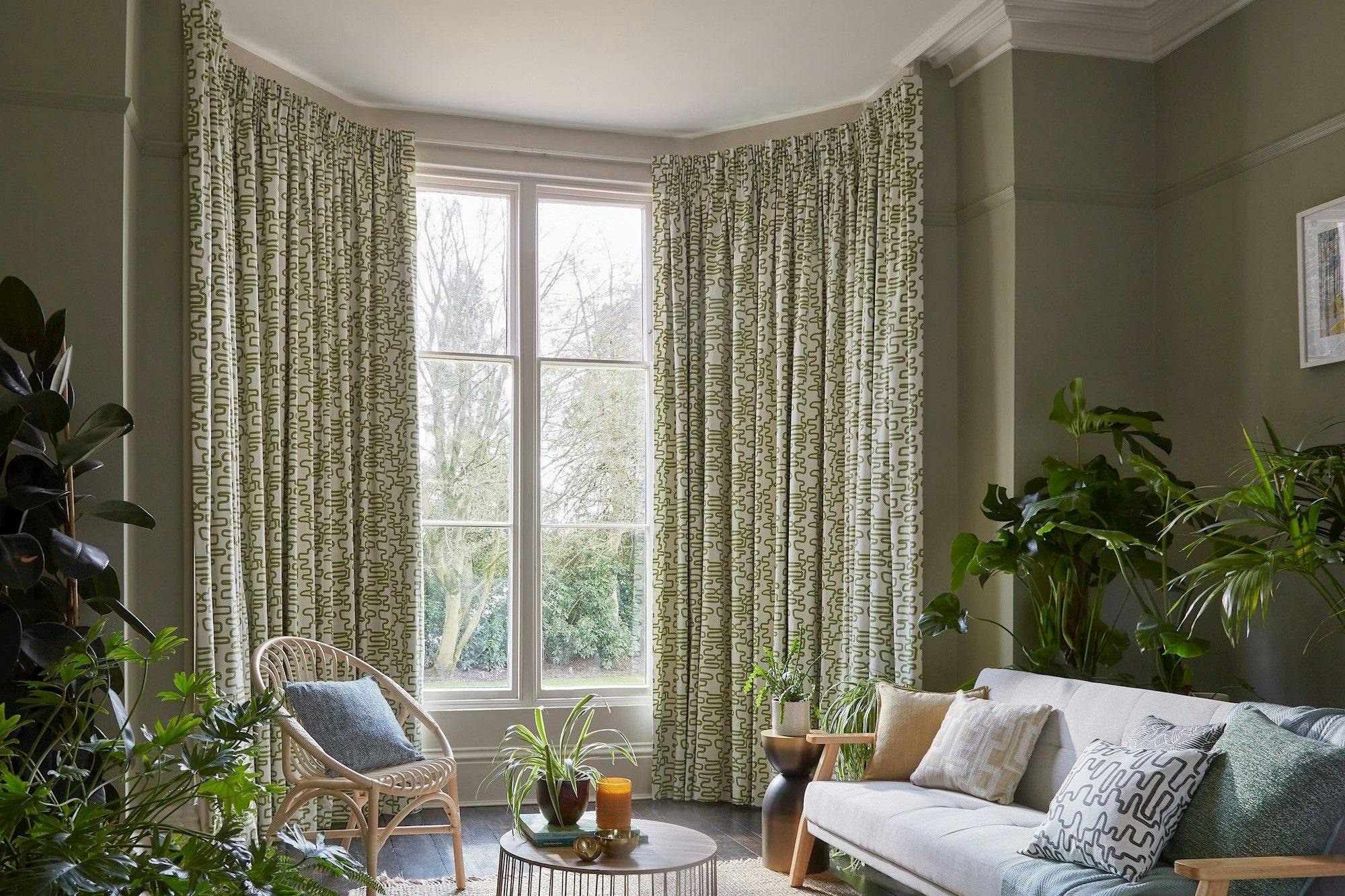 Curtains hang beside a large window in a living room, surrounding a wicker chair and a table with plants. A sofa with cushions is nearby, and plants decorate the space.