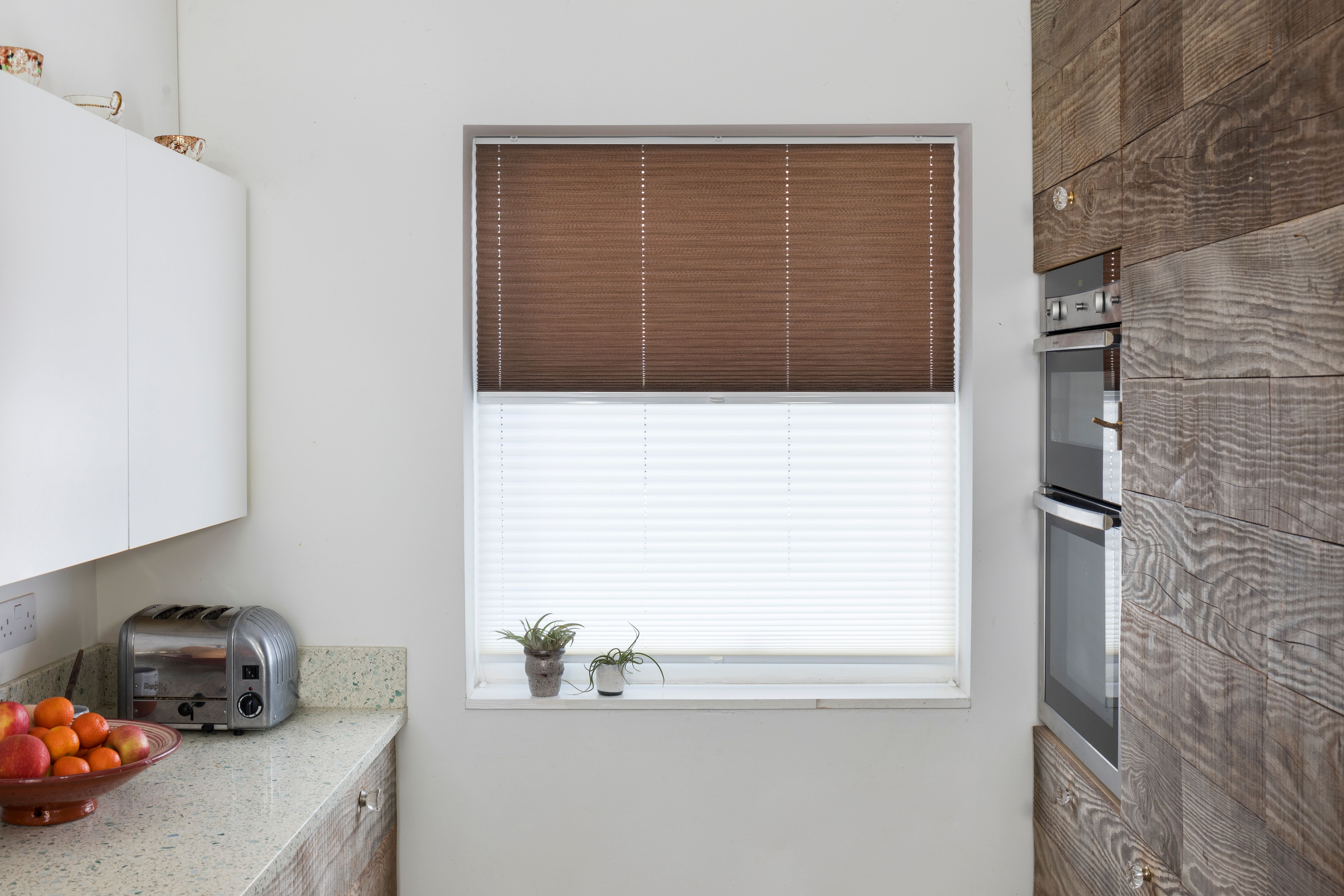 A kitchen window with brown blinds partially open, revealing a counter below with a toaster and fruit bowl, surrounded by white cabinets and wooden walls.