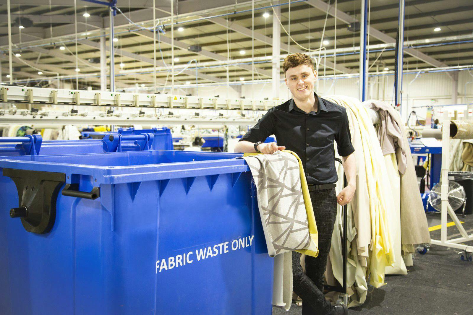 A person stands smiling beside a large blue bin labeled "FABRIC WASTE ONLY," holding fabric against a background of rolls of cloth and factory equipment.
