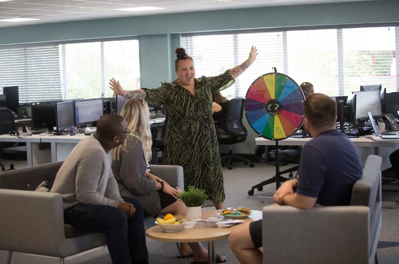 A woman enthusiastically gestures near a colorful spinning wheel in an office, while three seated colleagues watch. The room contains desks, computers, and large windows.