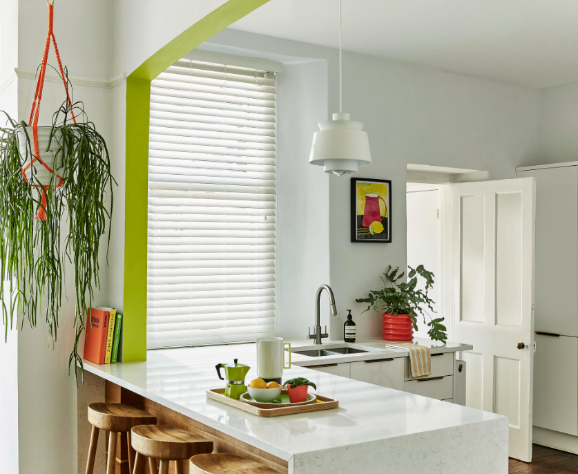A modern kitchen features a white island with wooden stools, topped with a tray of kitchenware, situated under a pendant light. Potted plants and colorful accents brighten the space.