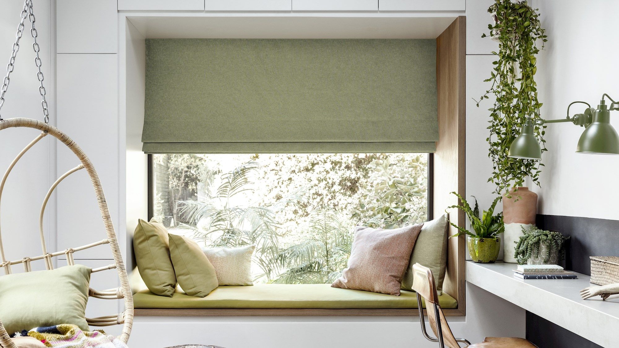 Window seat with green cushions overlooks plants through a large window with a green shade. Nearby, a swing chair and a desk with plants and books complete the cozy room.