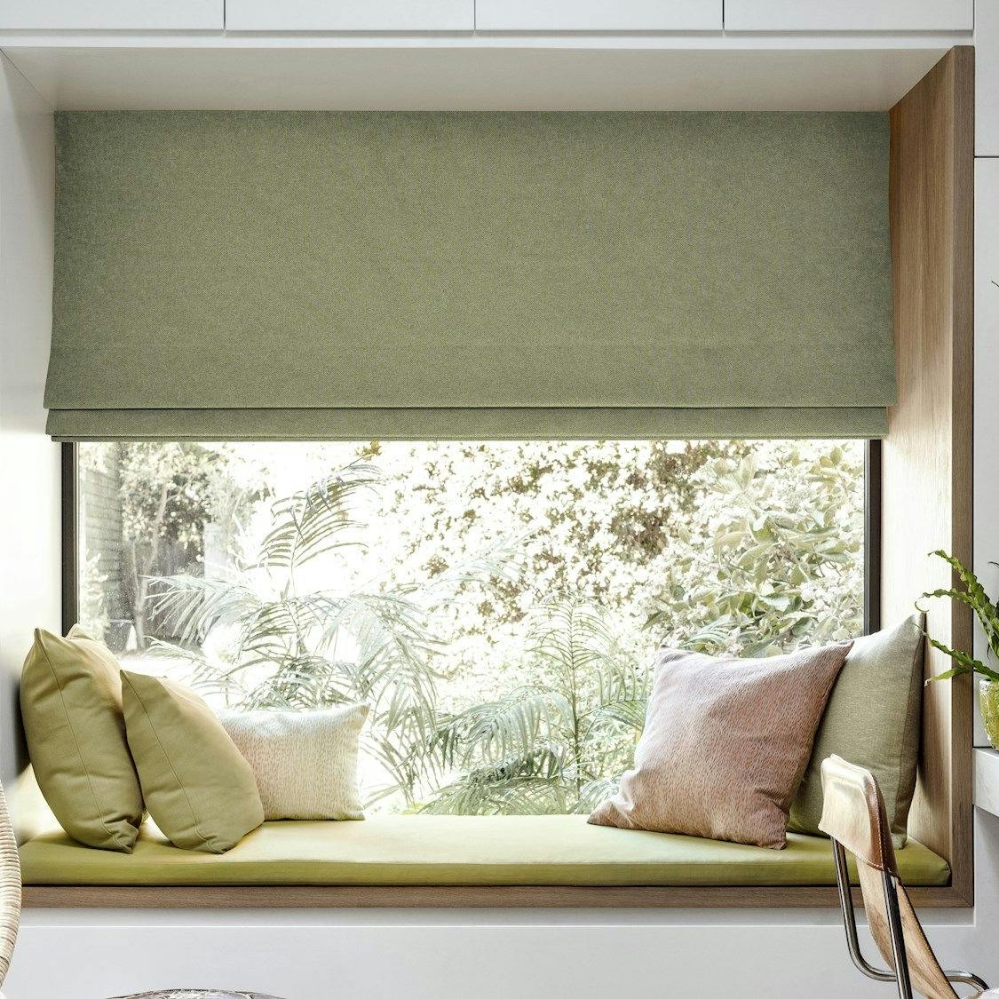 Window seat with green cushions overlooks plants through a large window with a green shade. Nearby, a swing chair and a desk with plants and books complete the cozy room.