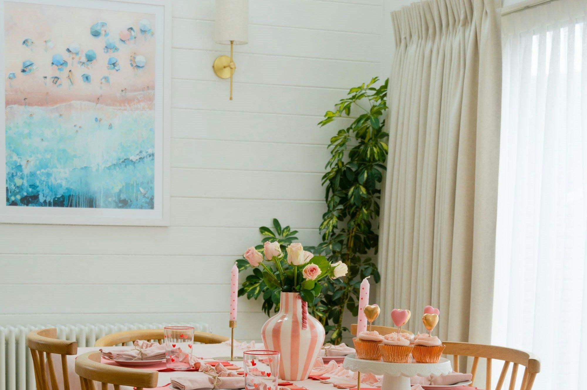 A dining table adorned with pink roses in a striped vase and heart-topped cupcakes, set amid a bright room with white walls, abstract artwork, and a leafy plant.