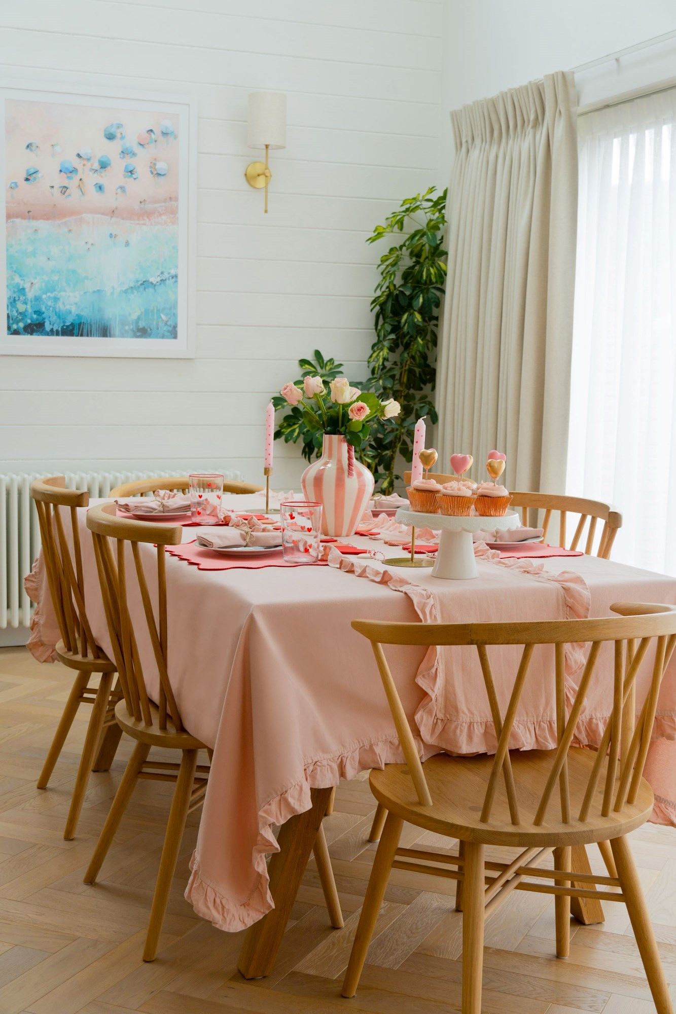 A dining table is adorned with a pink tablecloth and set for a meal, featuring cupcakes and a vase of roses. The room has wooden chairs, a bright painting, and a leafy plant.