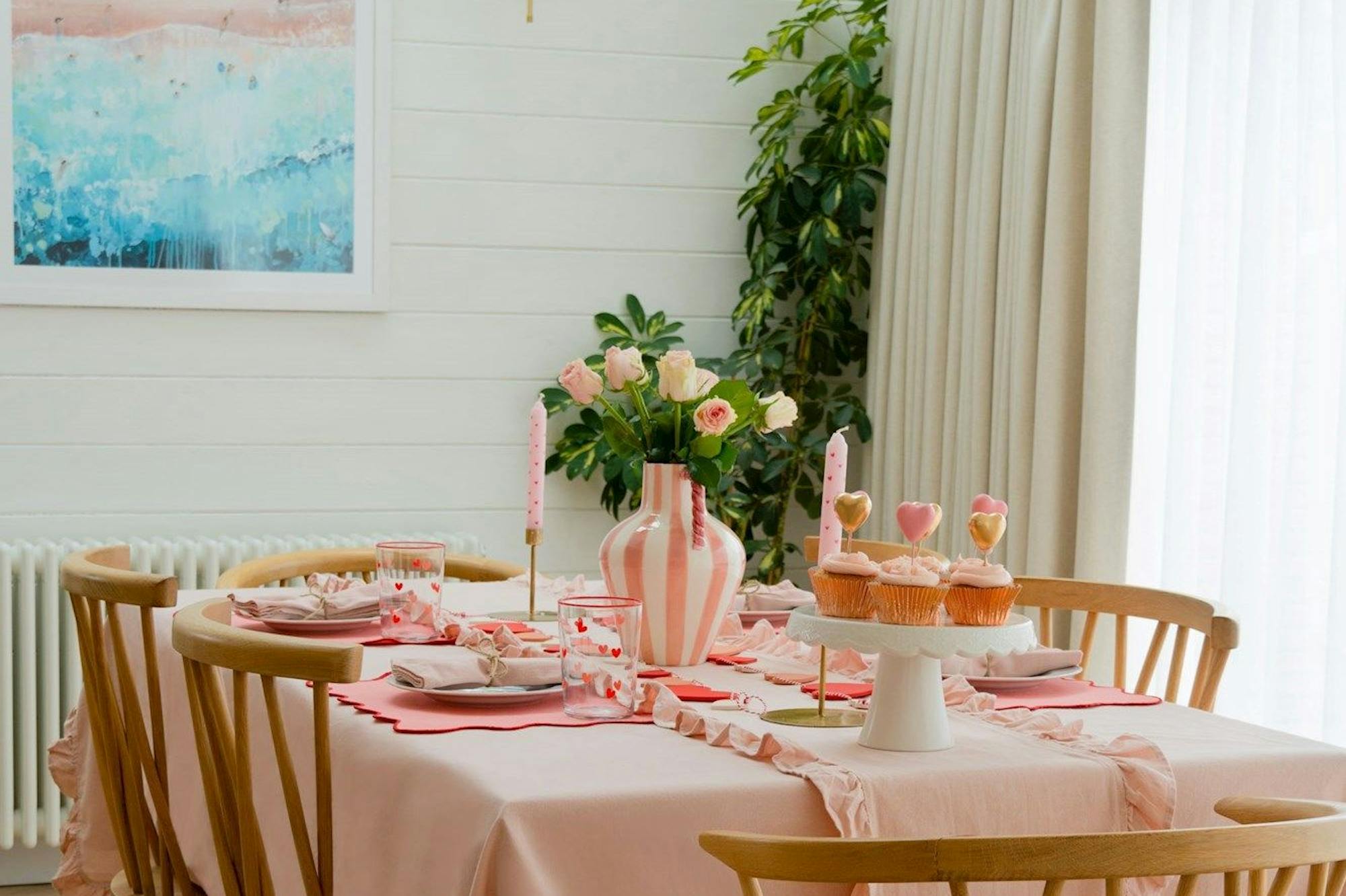 A dining table is adorned with a pink tablecloth and set for a meal, featuring cupcakes and a vase of roses. The room has wooden chairs, a bright painting, and a leafy plant.
