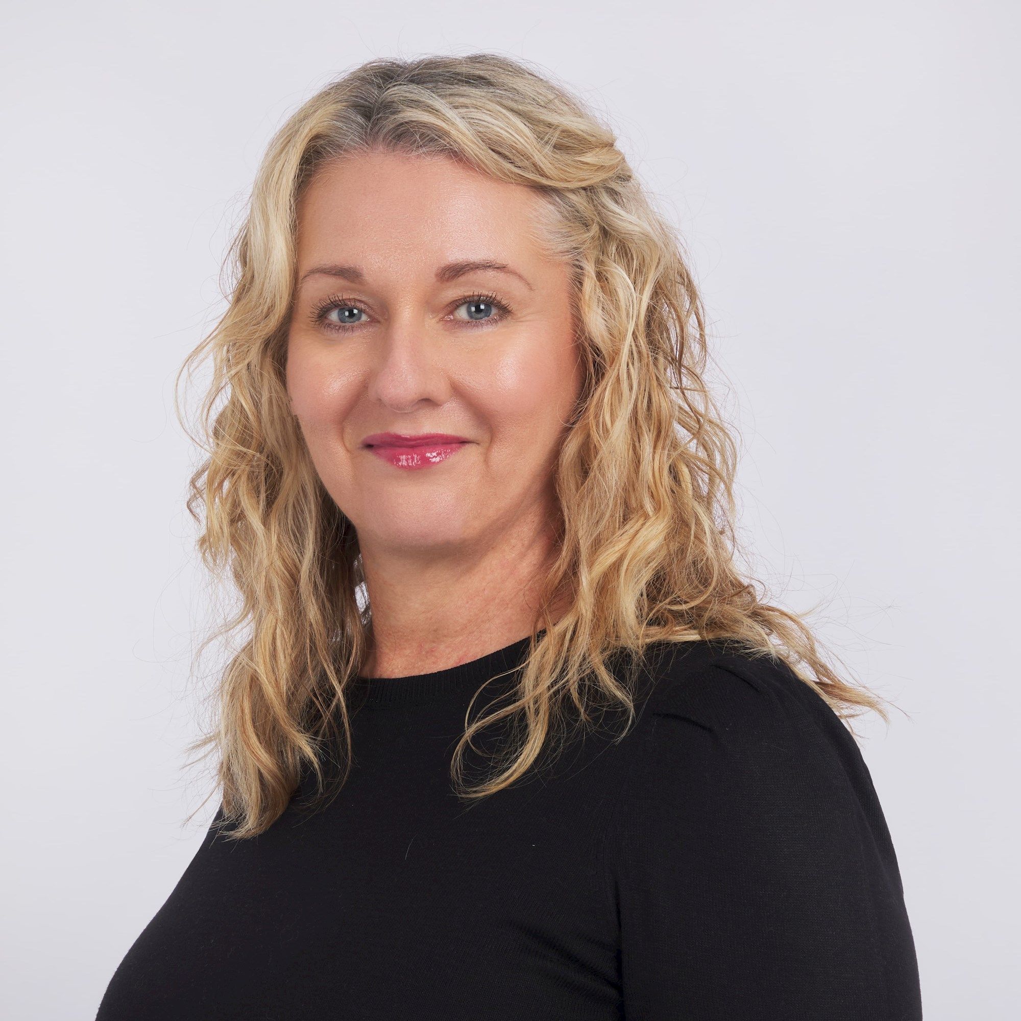 A person with wavy, blonde hair smiles slightly, wearing a black top. They are set against a plain, light background.