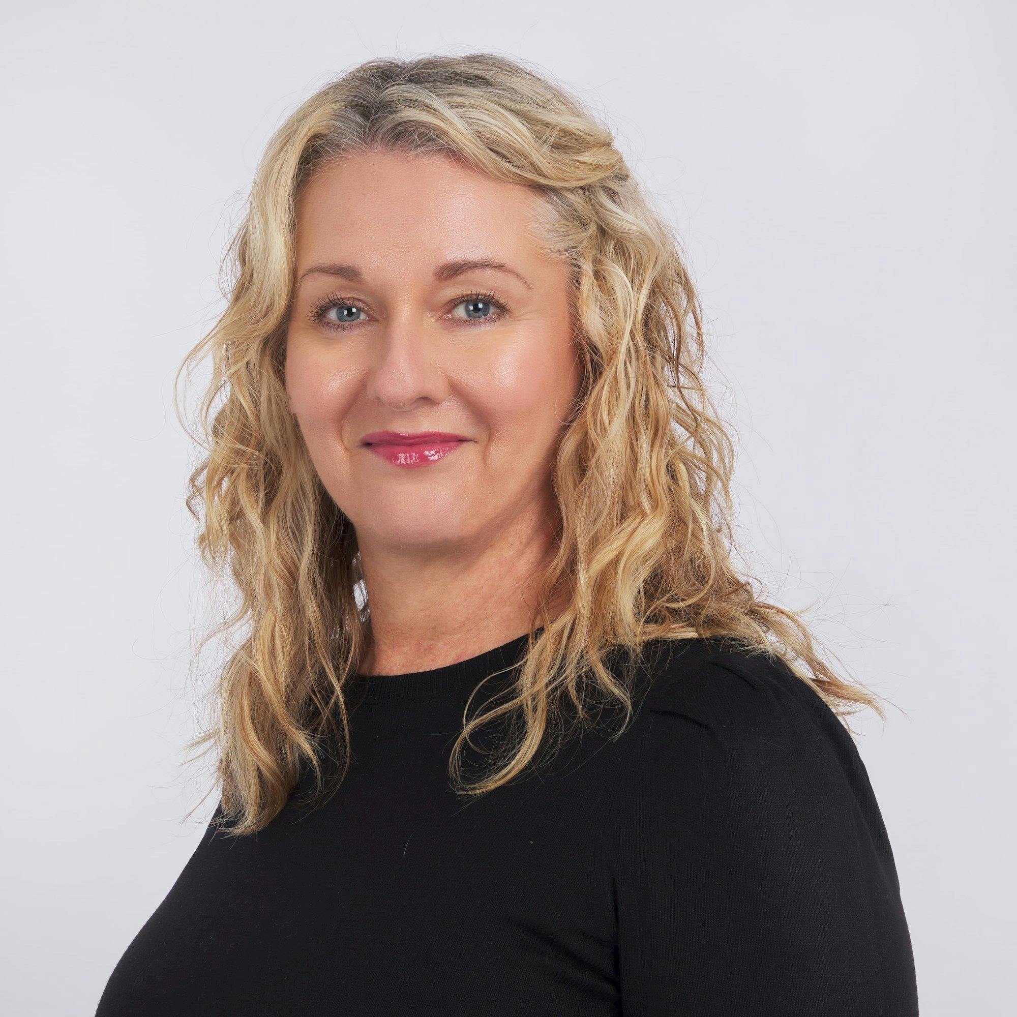 A person with wavy, blonde hair smiles slightly, wearing a black top. They are set against a plain, light background.