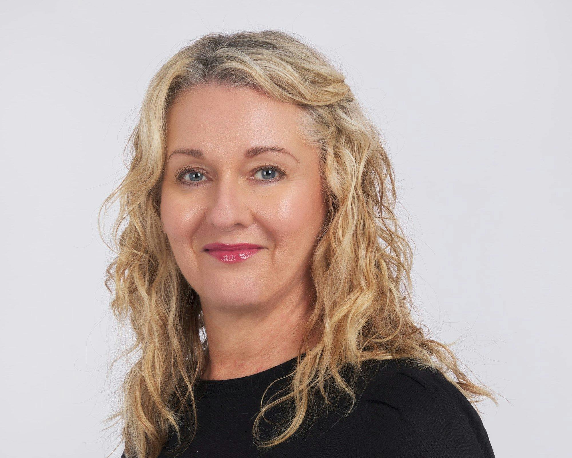 A person with wavy, blonde hair smiles slightly, wearing a black top. They are set against a plain, light background.