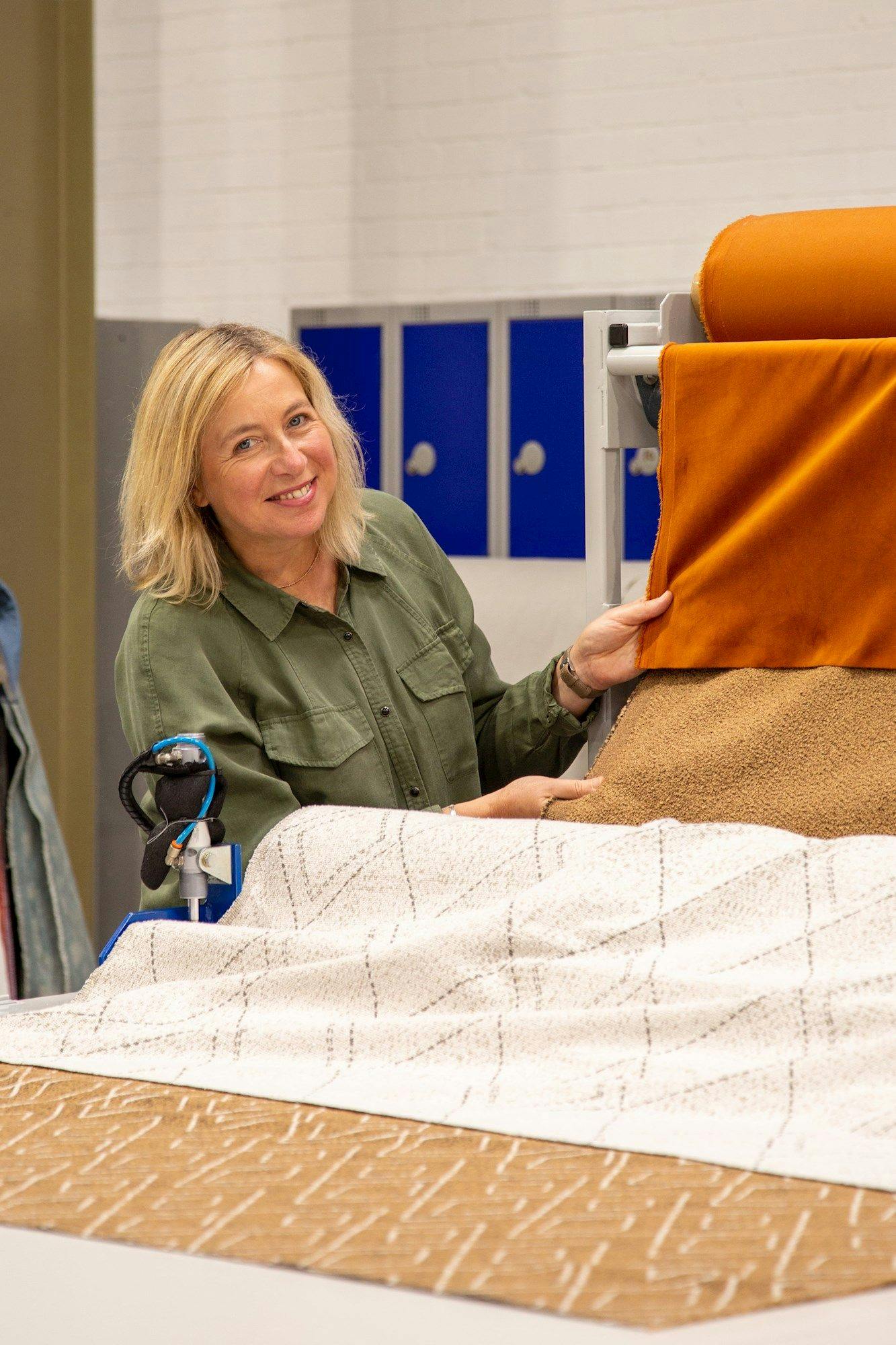 A person examines textile samples on a display rack, including orange velvet and beige fabric, in a workshop with blue lockers and white brick walls.