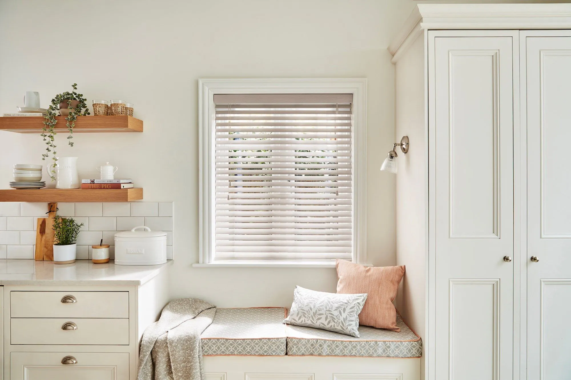 A built-in window seat between kitchen cabinets painted in warm white. The seat is topped with cushions in neutral shades and the window is dressed with a white wooden Venetian blind.  