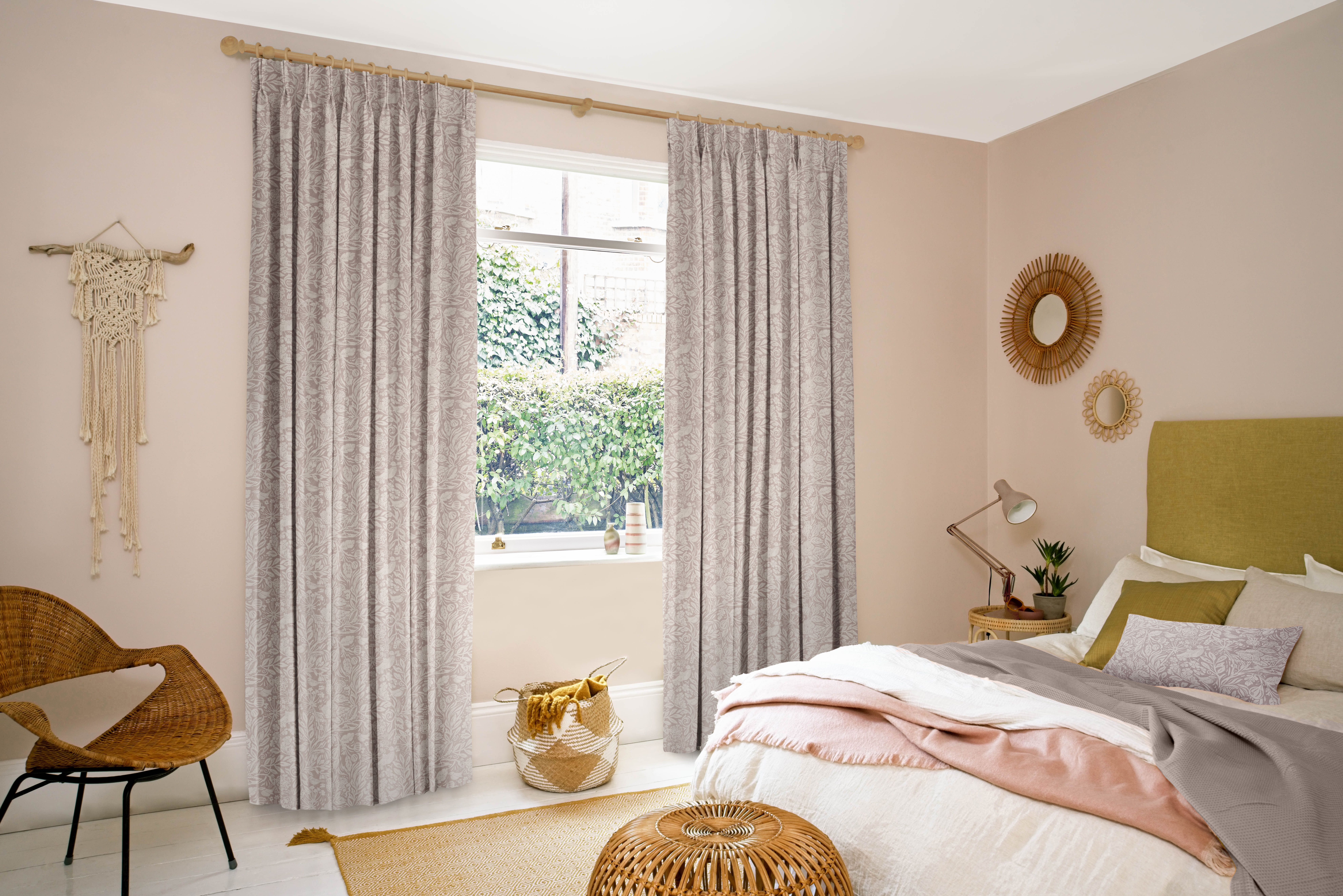 Curtains hang beside a window adorned with greenery, in a cozy bedroom with a bed, wicker chair, and decorative items. Soft light creates a serene, inviting atmosphere.
