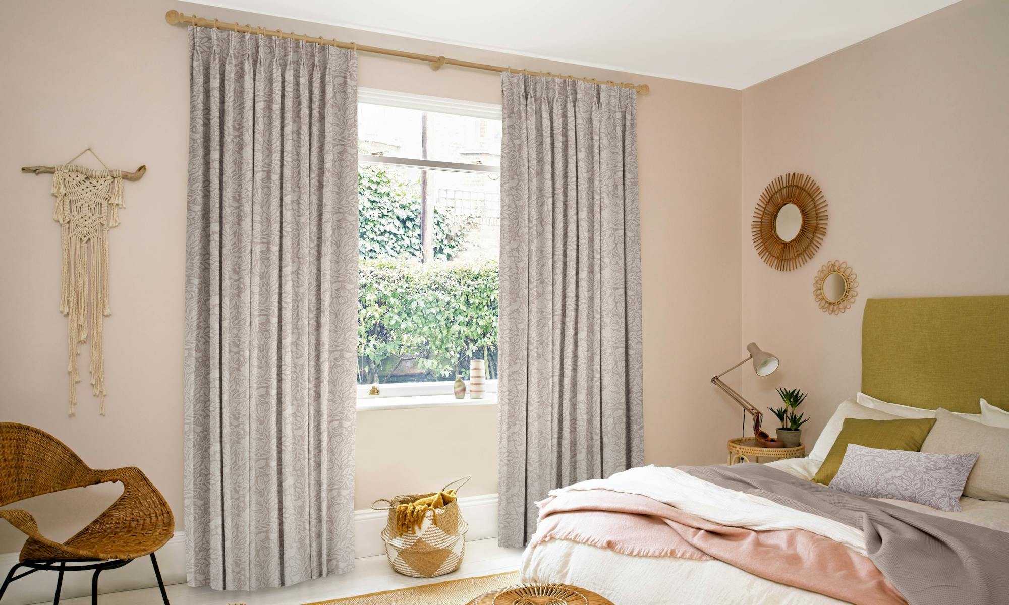 Curtains hang beside a window adorned with greenery, in a cozy bedroom with a bed, wicker chair, and decorative items. Soft light creates a serene, inviting atmosphere.