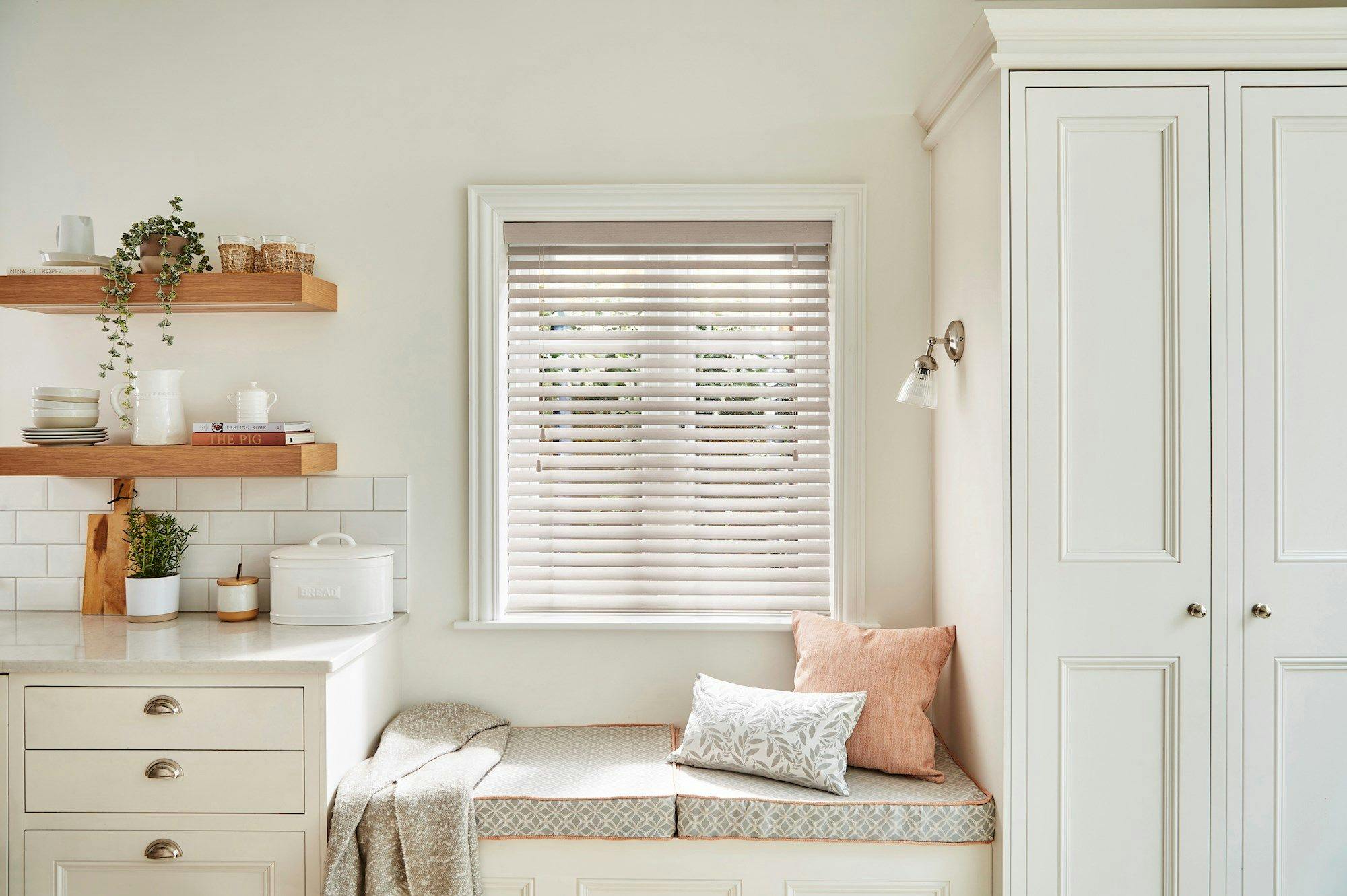 A cozy kitchen nook with a bench adorned with cushions sits under a window with beige blinds. Nearby, shelves hold plants, dishes, and books.