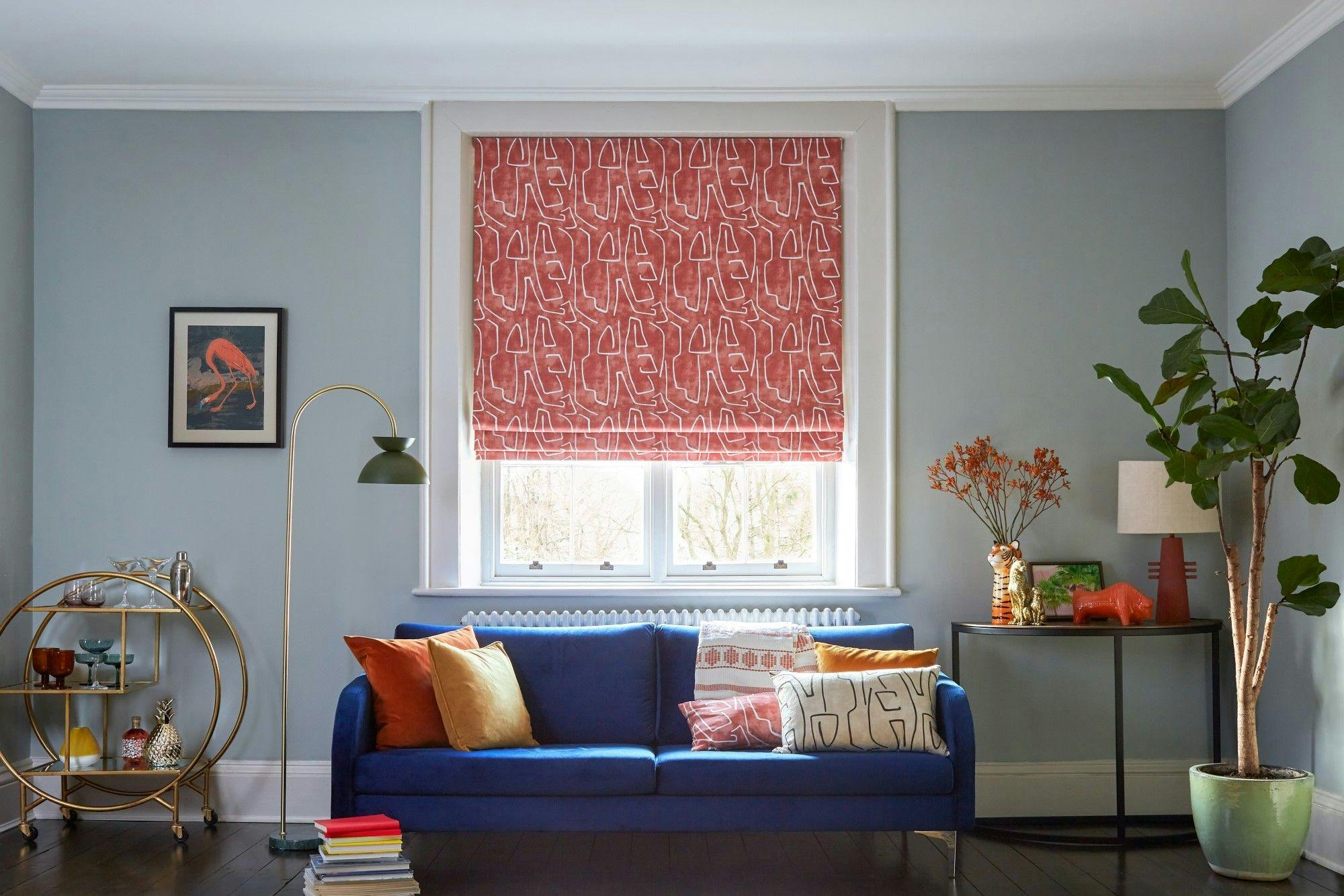 A blue sofa with colorful cushions sits under a large window with patterned red blinds. A bar cart, floor lamp, and plant adorn the stylish, light blue room.