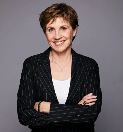 A person with short hair smiles while standing with arms crossed, wearing a pinstripe suit over a white shirt, set against a plain gray background.
