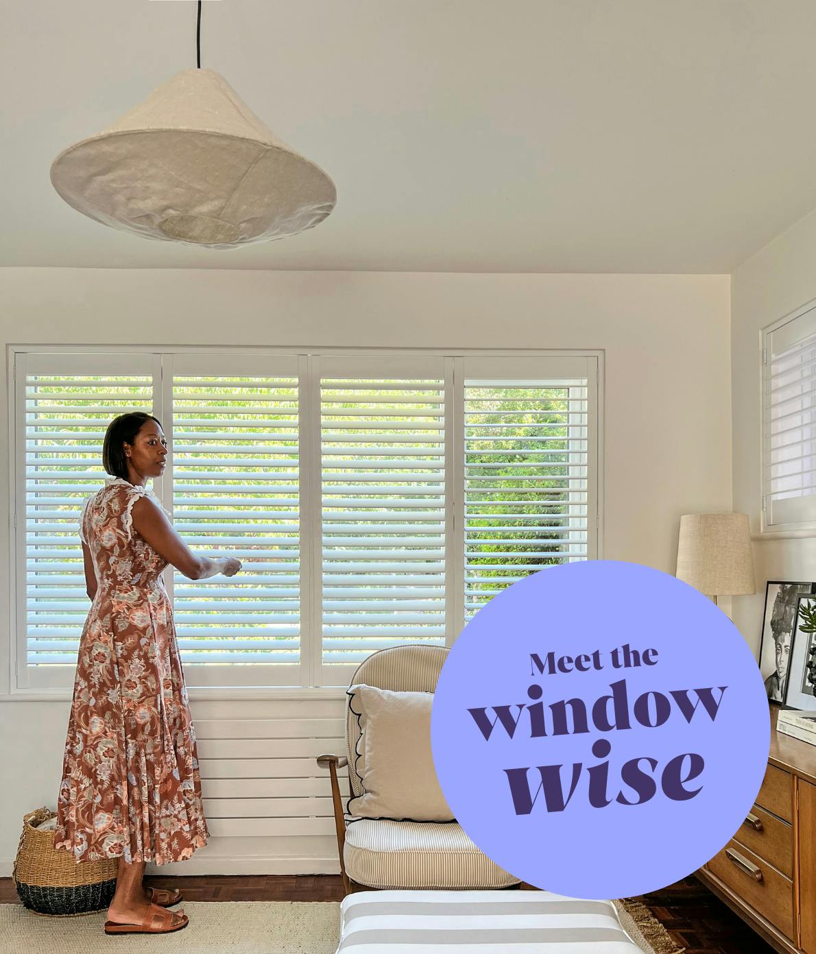 A person in a floral dress adjusts window blinds in a cozy room with a chair, lamp, and cabinet. Text reads, "Meet the window wise" on a lavender circle.